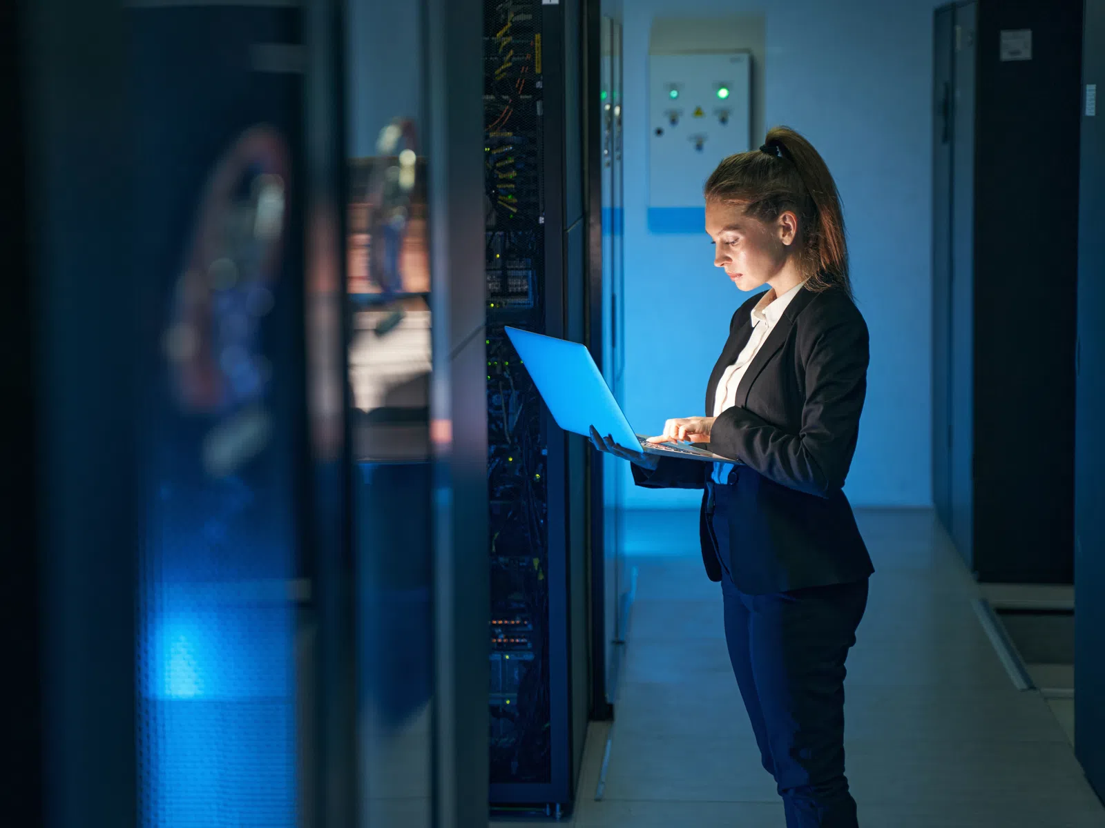 Woman in business attire working on a laptop in a server room with blue lighting and technology racks.