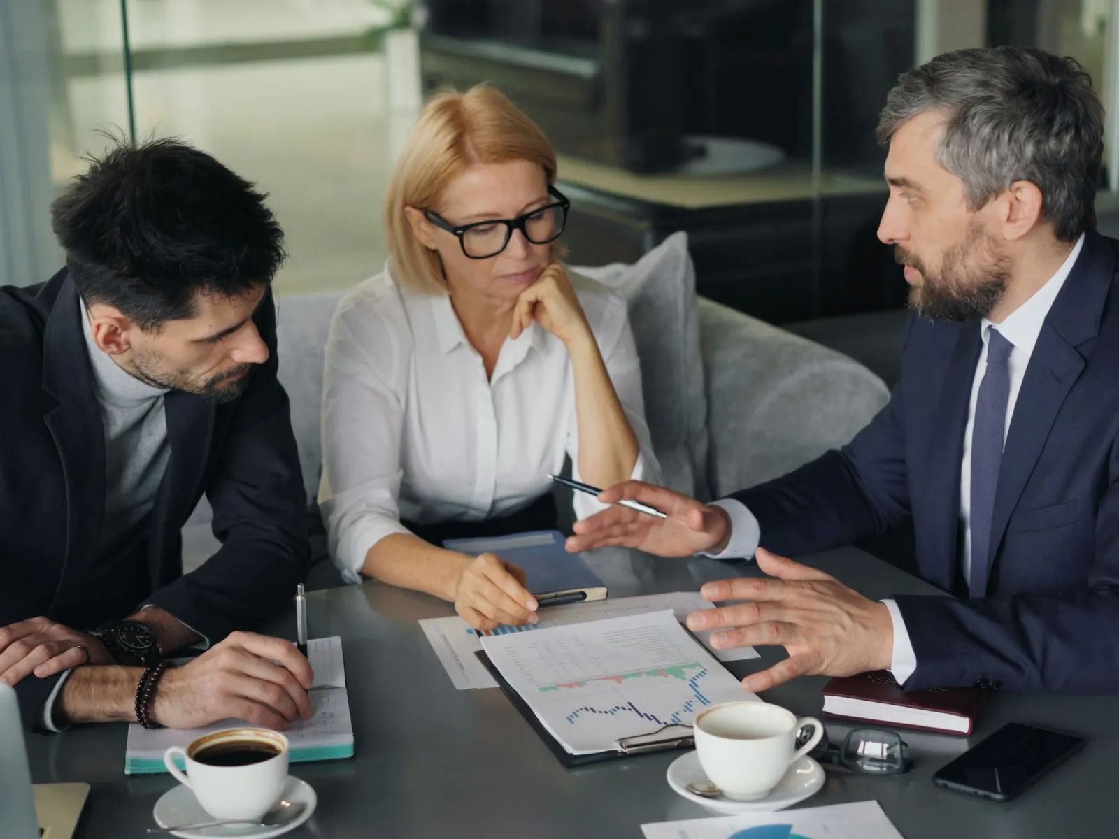 Three business professionals discussing charts and notes during a meeting in a modern office setting.
