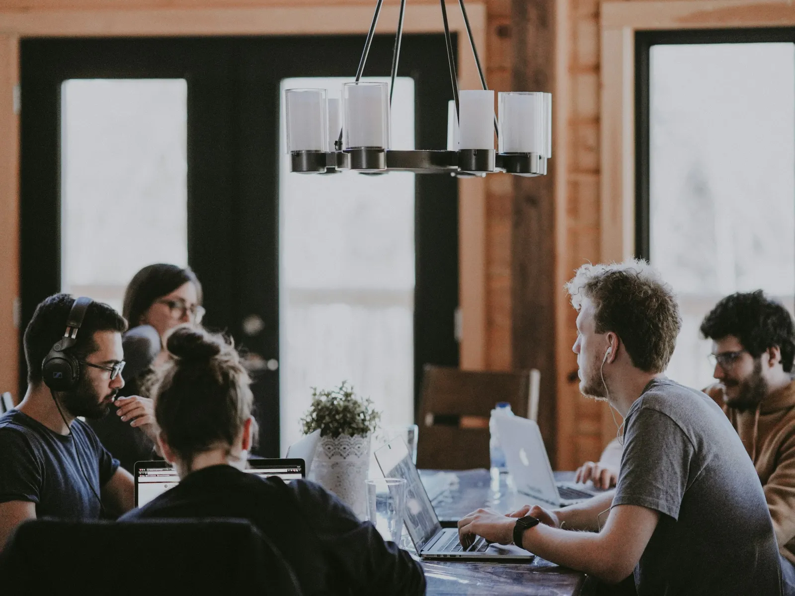 Group of young professionals collaborating with laptops in a cozy wooden room with large windows and modern lighting.