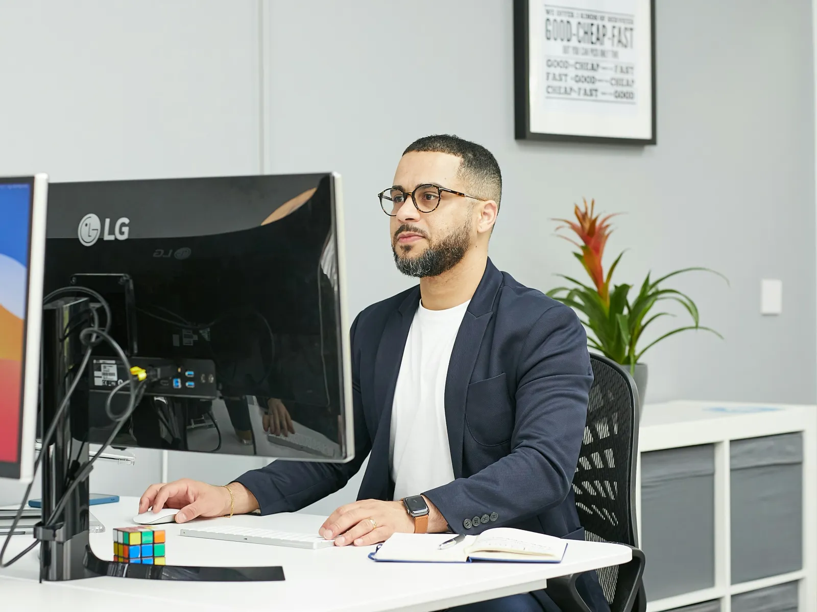 Man wearing glasses and blazer working on a computer at a modern office desk with a notebook and plant.
