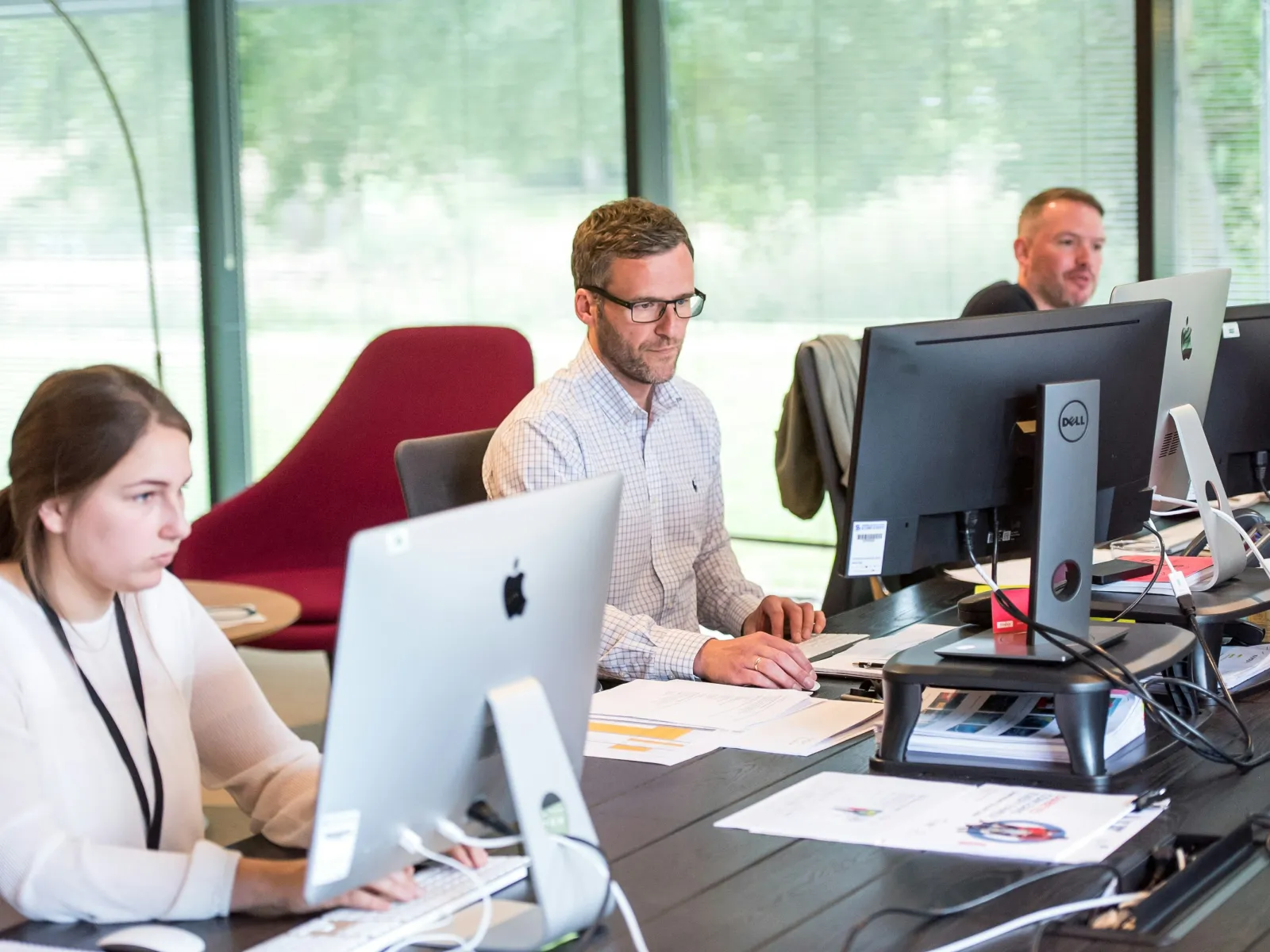 Three coworkers focused on their desktop computers in a bright modern office with large windows and a red chair.