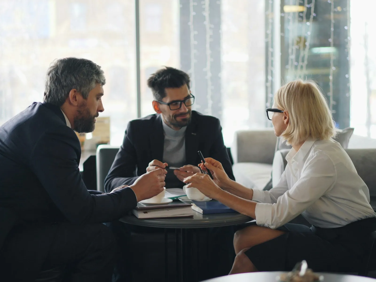 Three professionals in business attire discussing documents around a round table in a modern office setting.