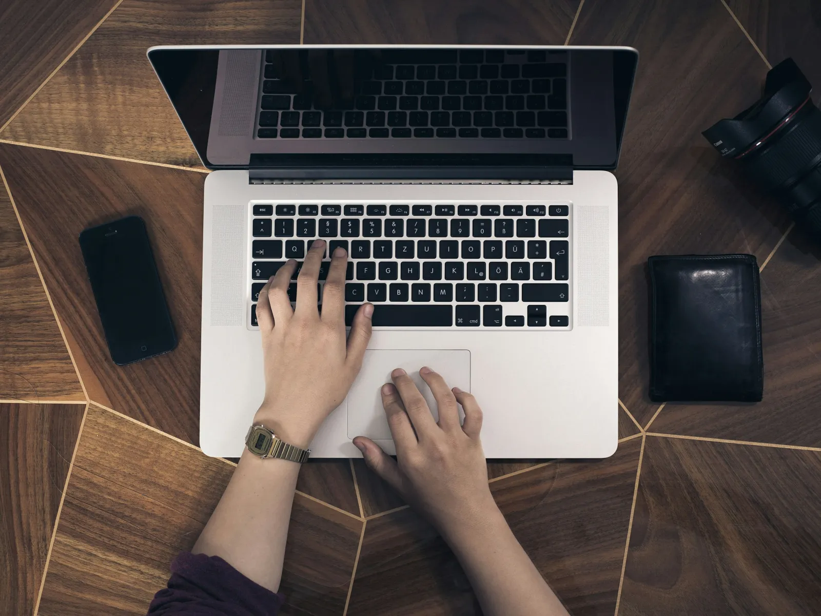 Person typing on a laptop keyboard on a wooden table with phone, camera lens, and wallet nearby
