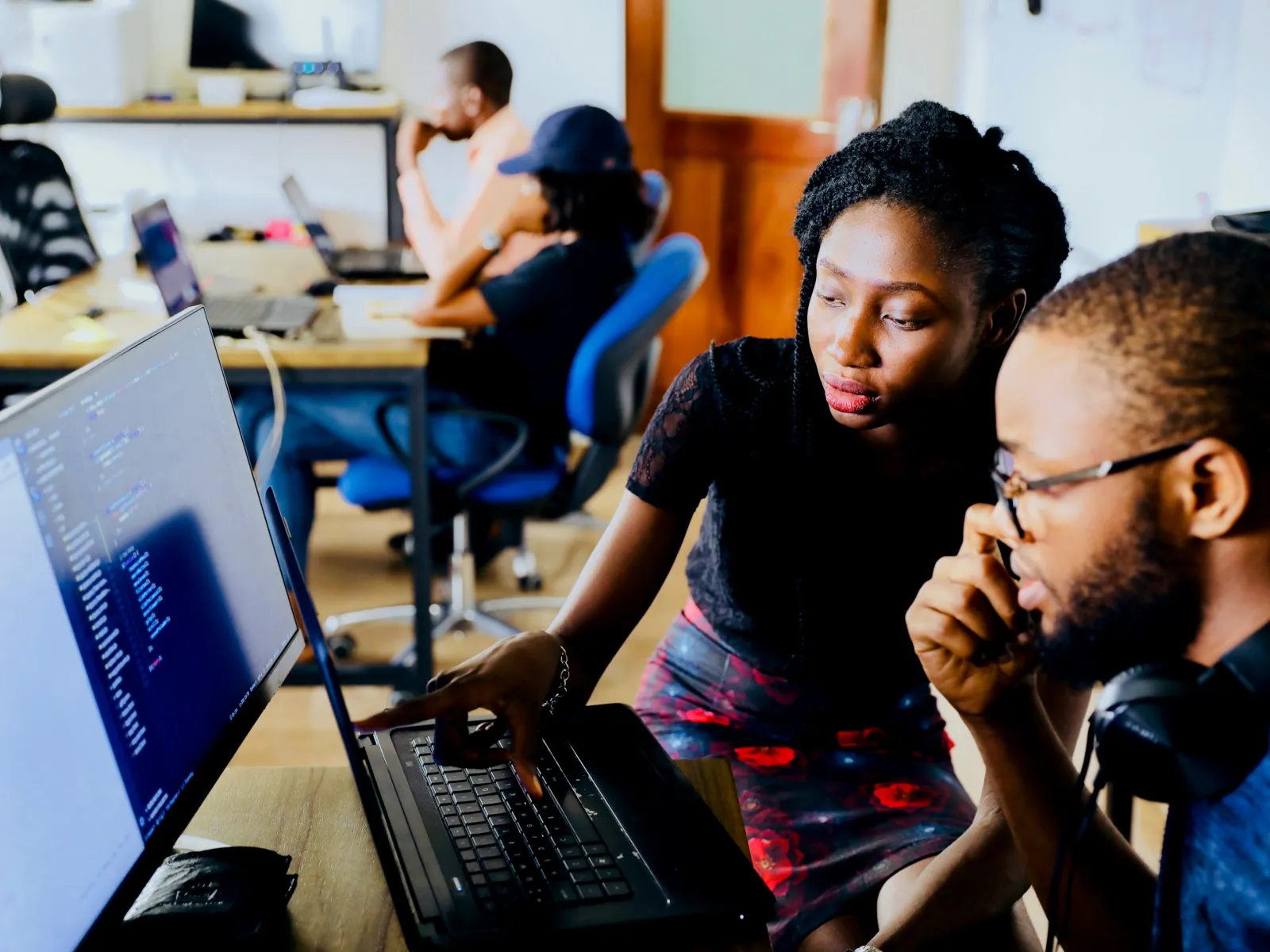 Two colleagues collaborating at a computer in a tech workspace with others working nearby.