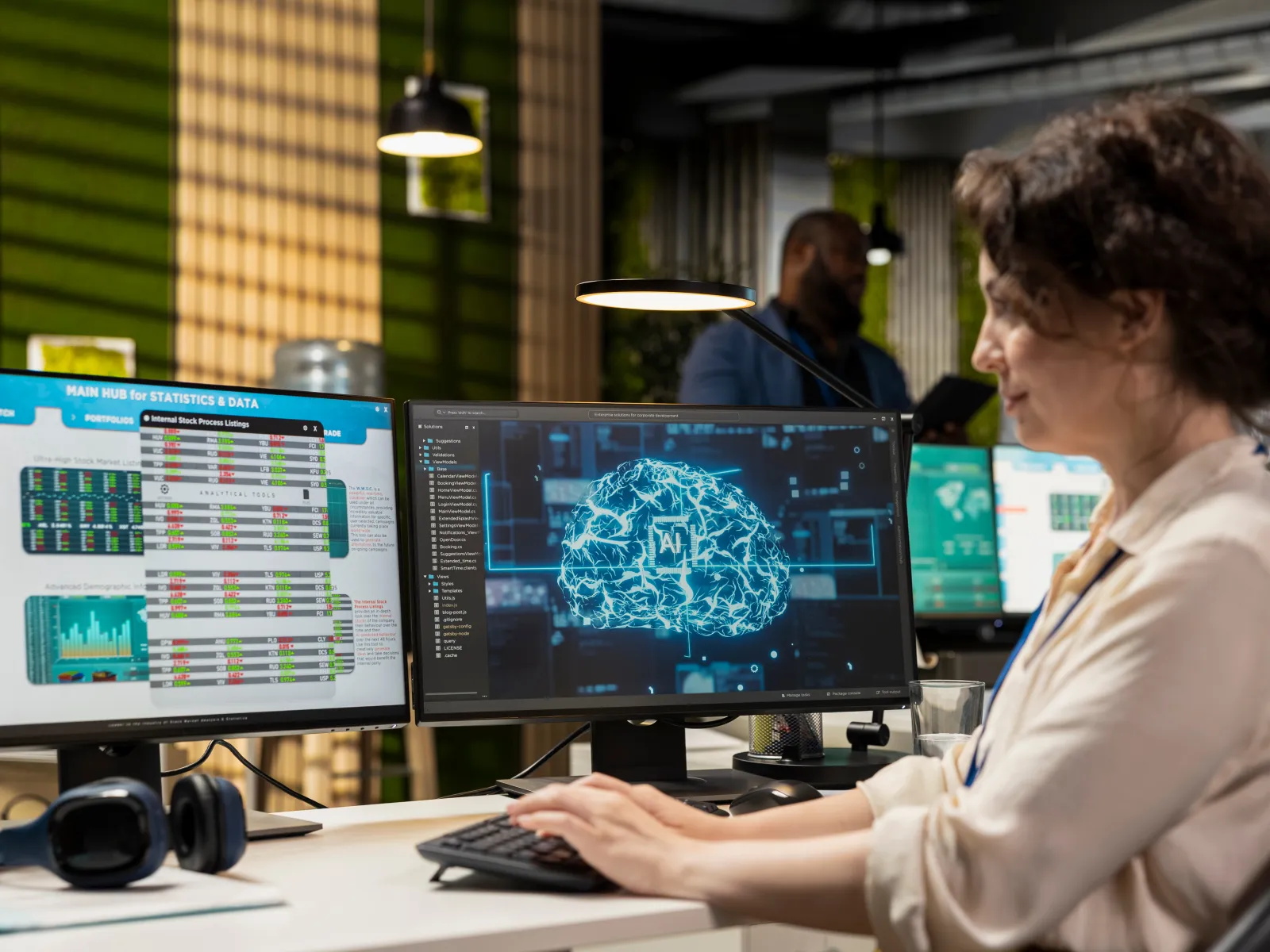 Woman working on AI brain visualization and data charts on dual monitors in a modern tech office.