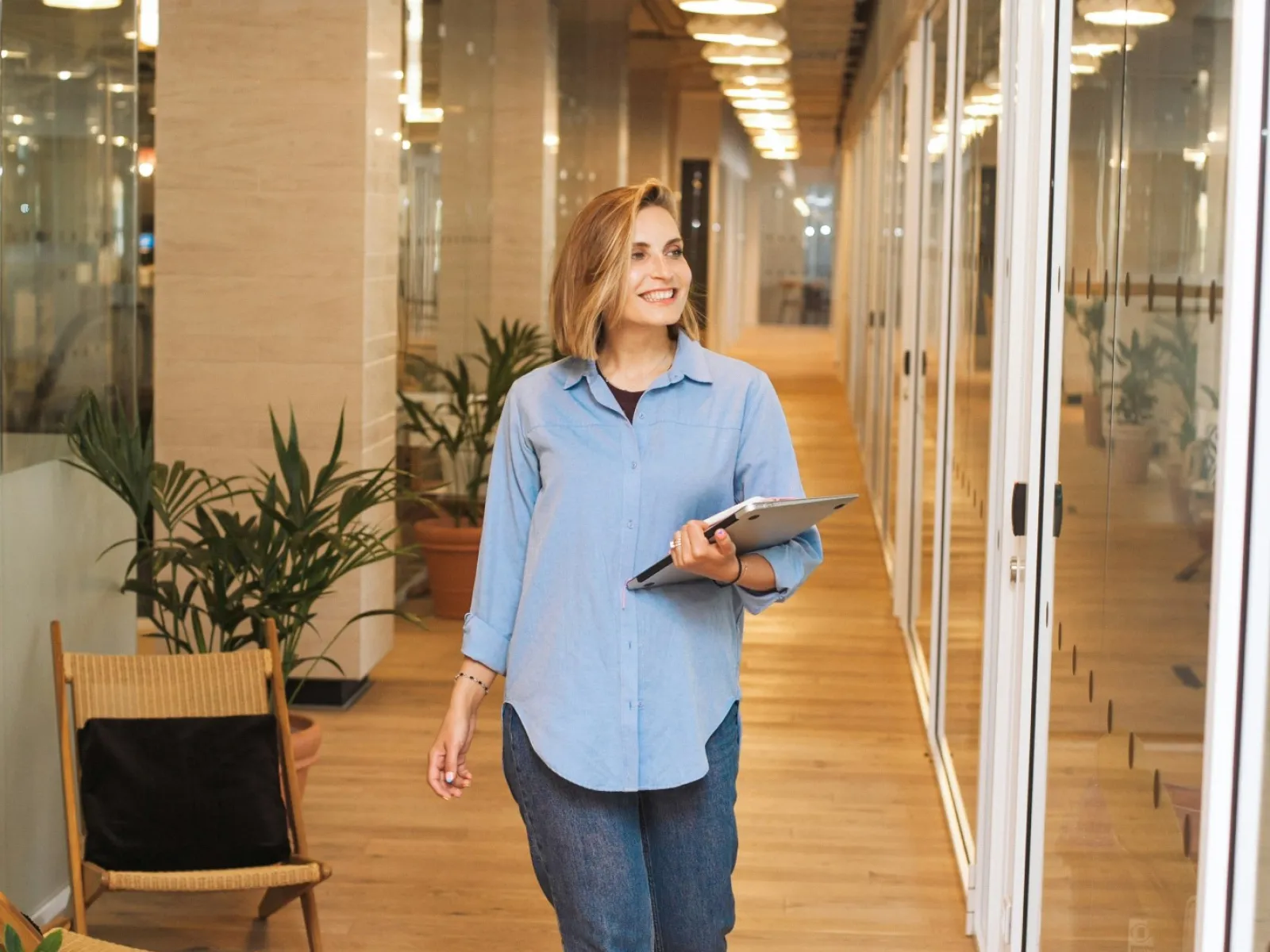 Smiling woman walking in modern office hallway holding a tablet and wearing casual clothes.