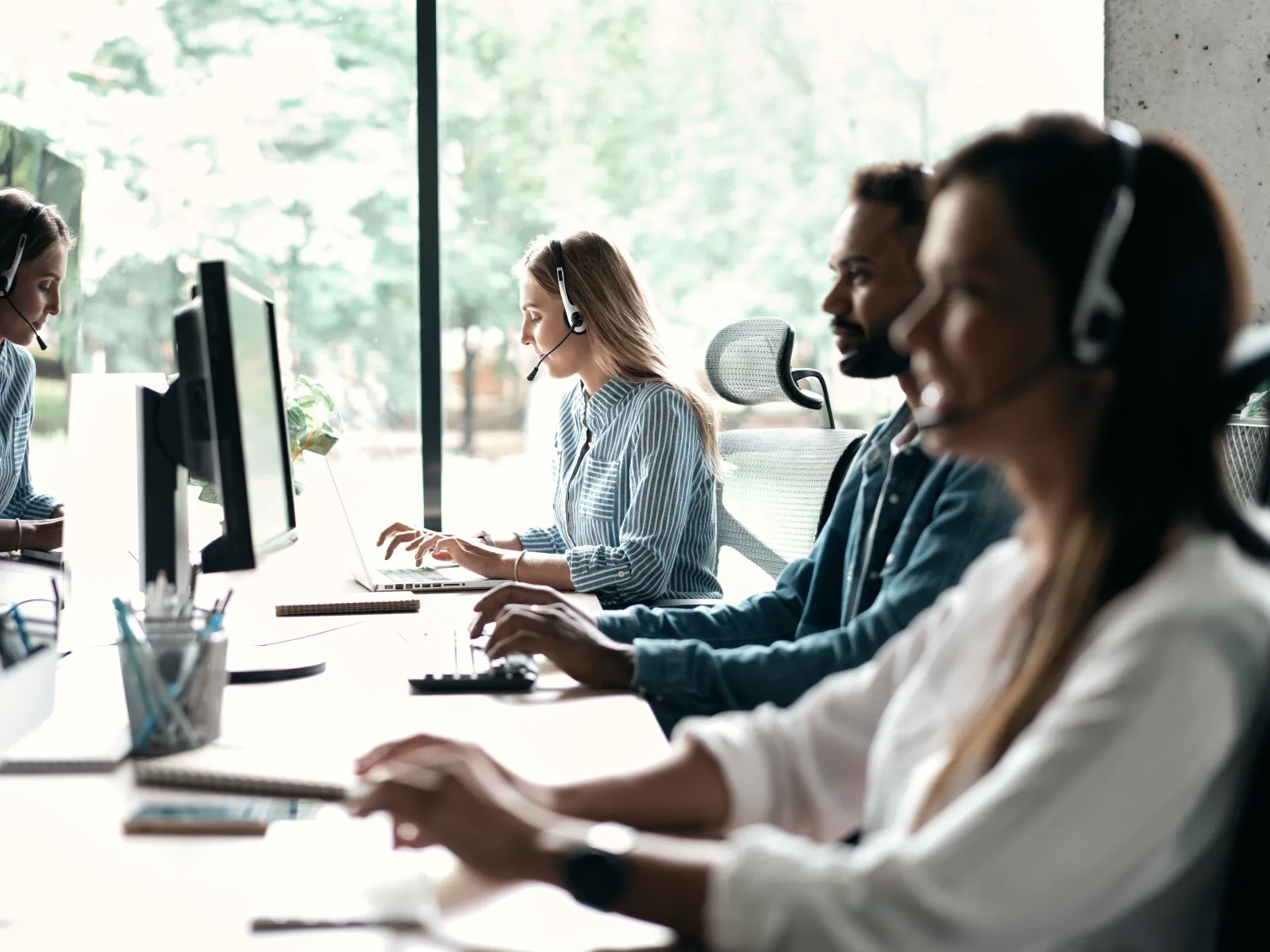 Customer service team working at computers wearing headsets in a bright modern office with large windows.