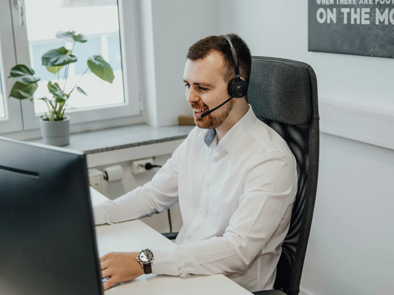 Smiling man in white shirt wearing headset working at computer in modern office with plant and window.