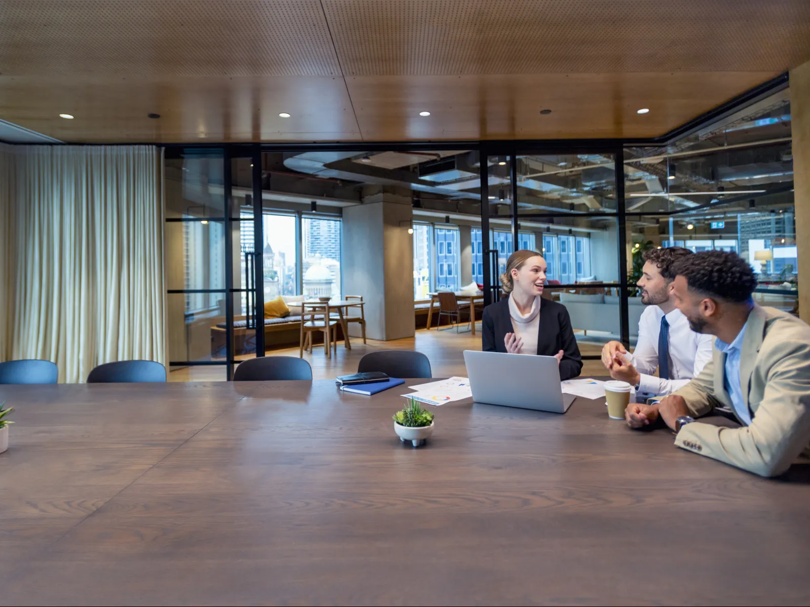 Three professionals engaged in discussion around a large wooden conference table in a modern office meeting room.