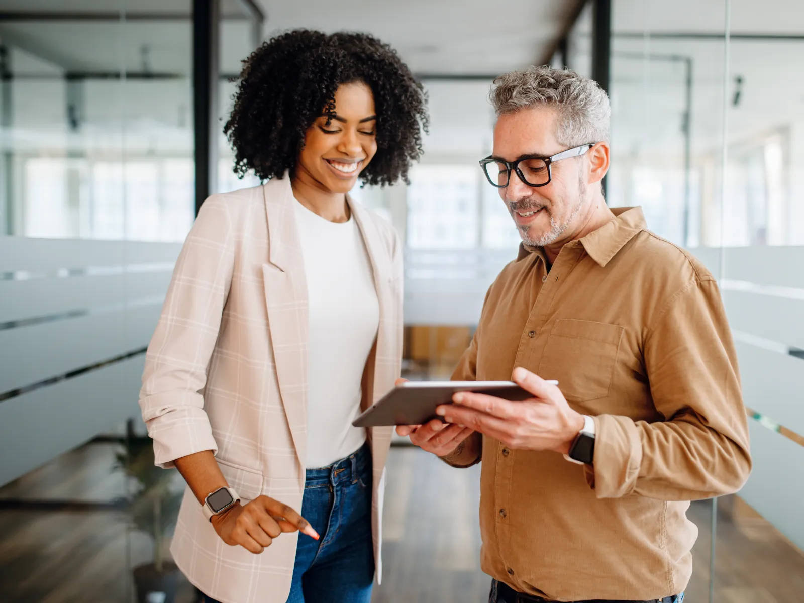 Two professionals smiling and discussing work using a digital tablet in a modern office corridor