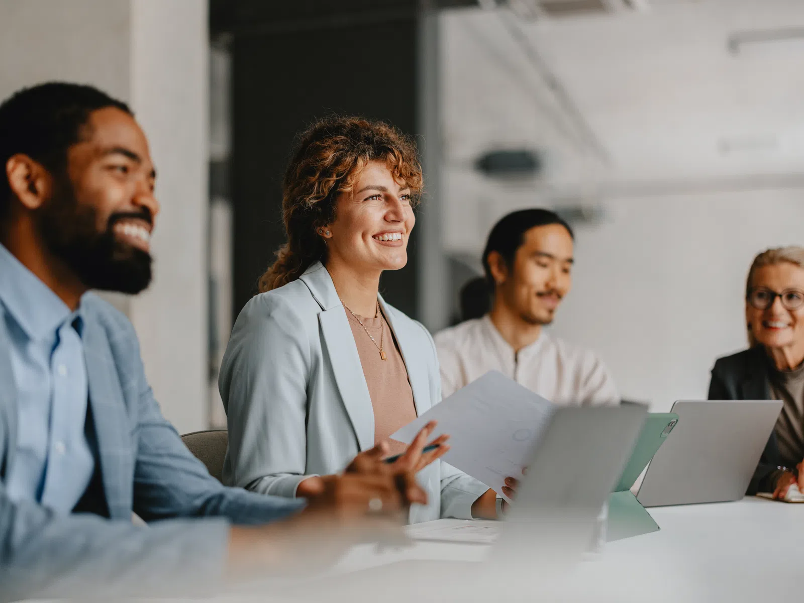 Diverse group of professionals smiling and collaborating in a modern office meeting around a table with laptops and documents.