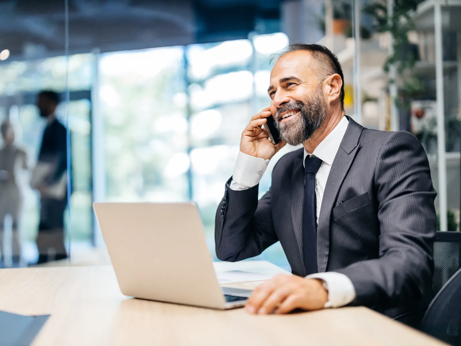 Smiling businessman in suit talking on phone while working on laptop in modern office setting