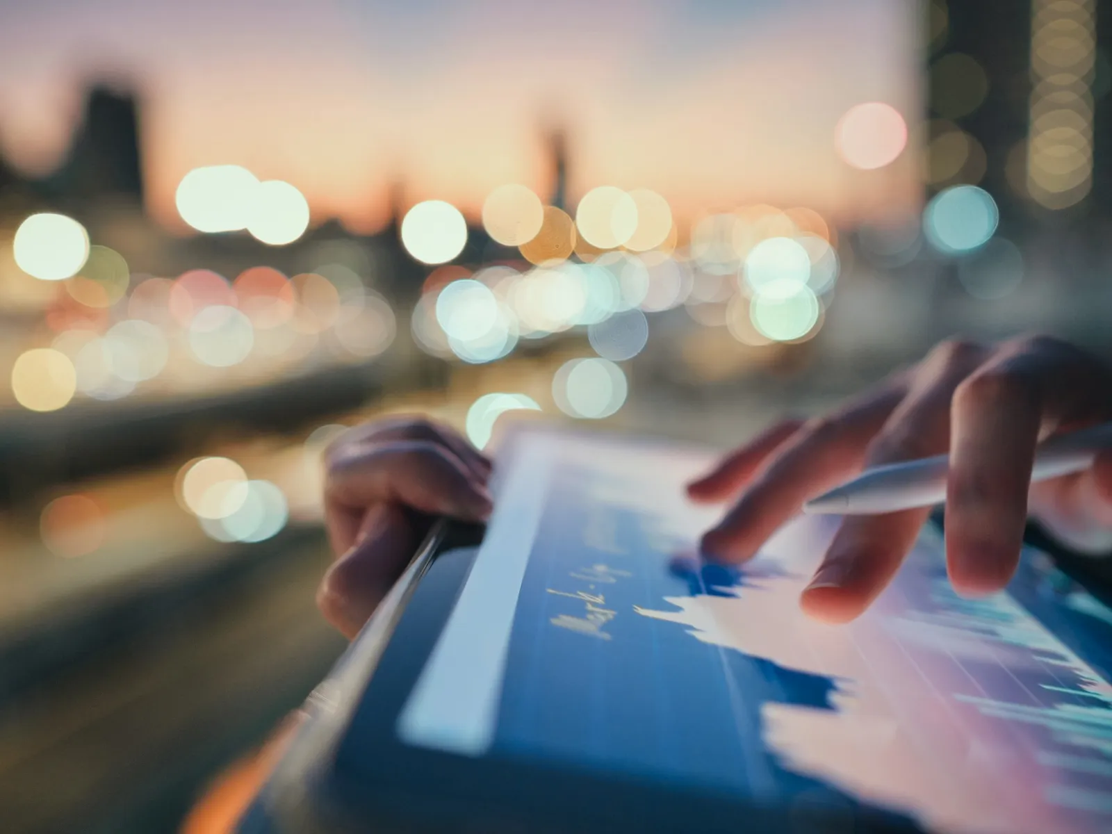 Person using stylus on tablet displaying financial charts with city lights blurred in the background at dusk