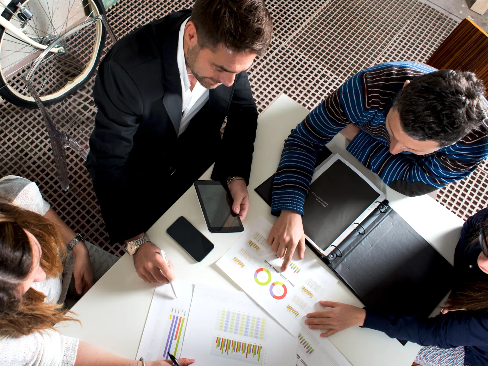 Overhead view of four people analyzing charts and graphs during a business meeting at a white table.