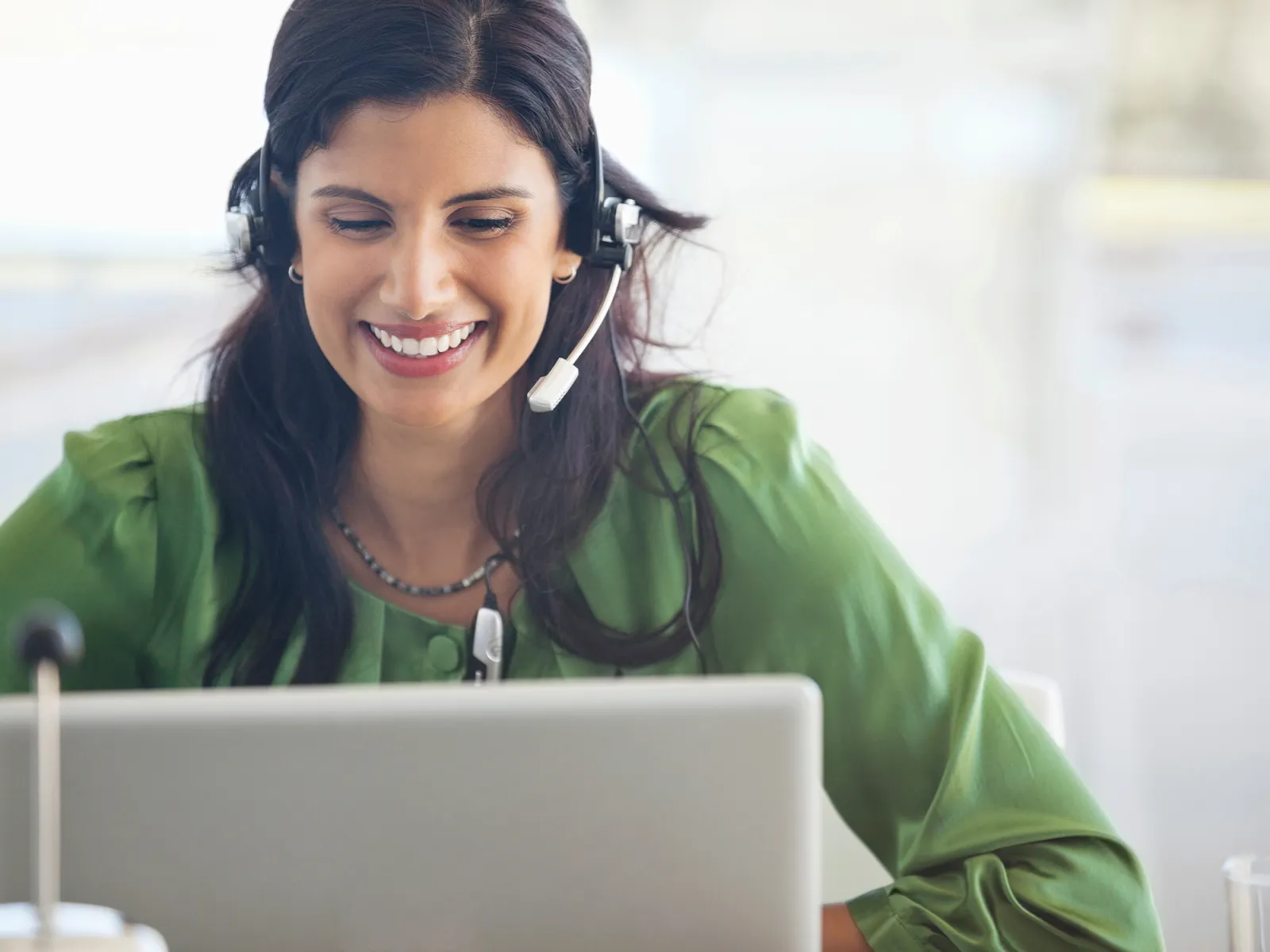 Smiling woman in green blouse wearing headset working on laptop in a bright office setting.