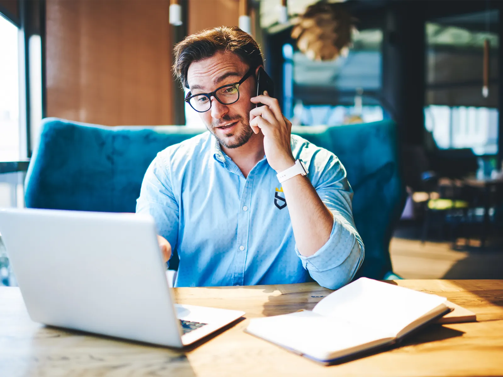 Man in blue shirt talking on phone and working on laptop at wooden desk with notebook in modern office.