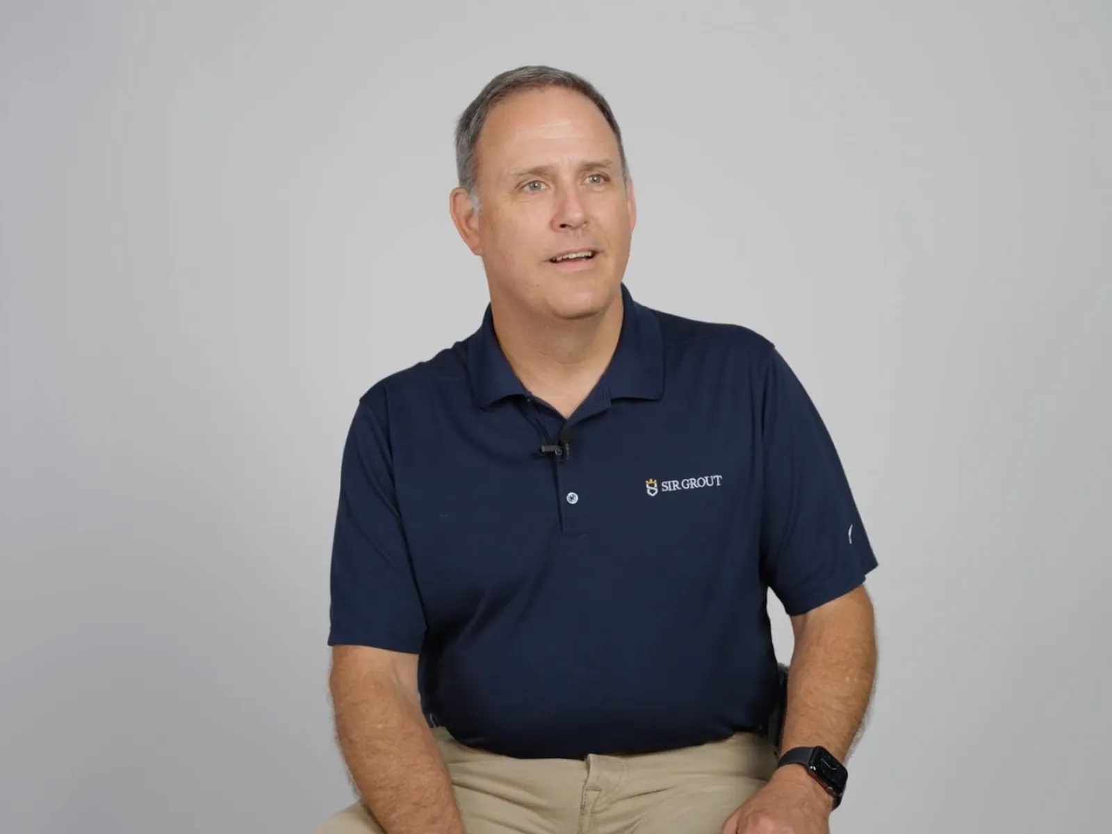 Middle-aged man in navy polo shirt and khaki pants sitting against a plain light gray background.