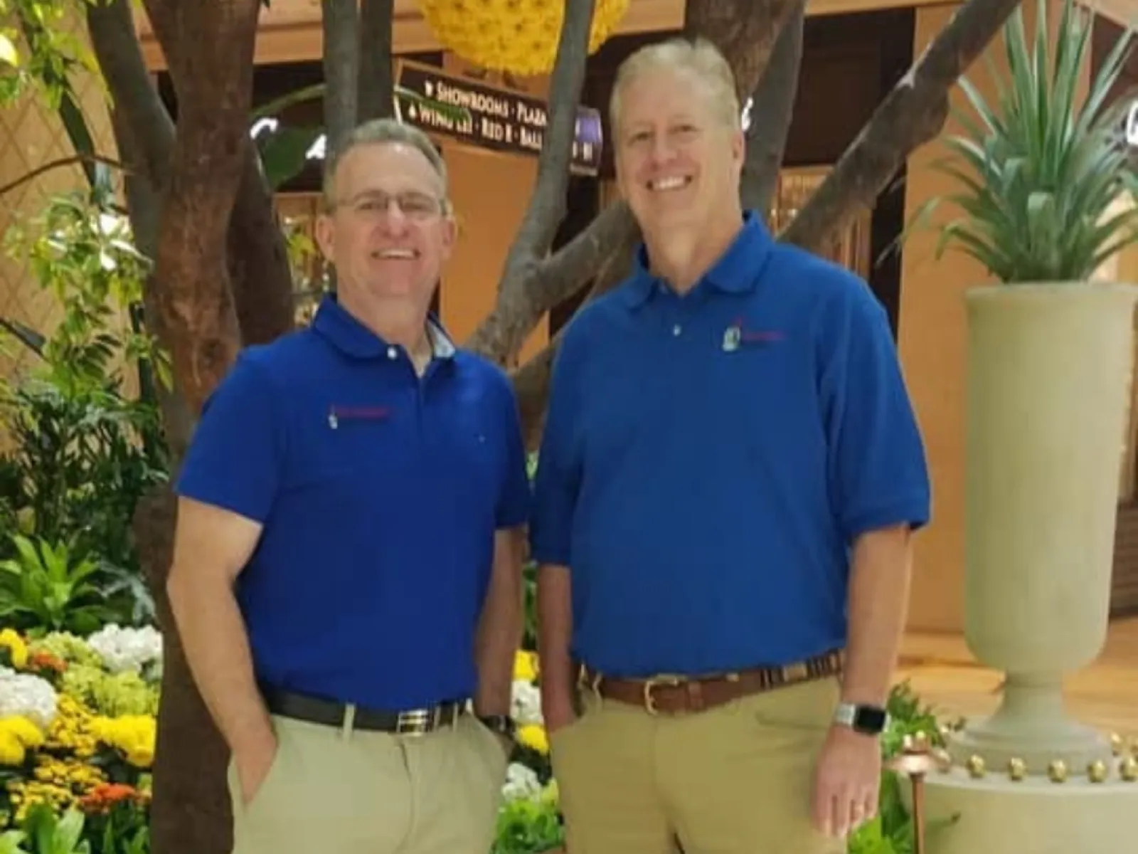 Two men in blue polo shirts and khaki pants standing indoors near plants and flowers smiling at the camera.