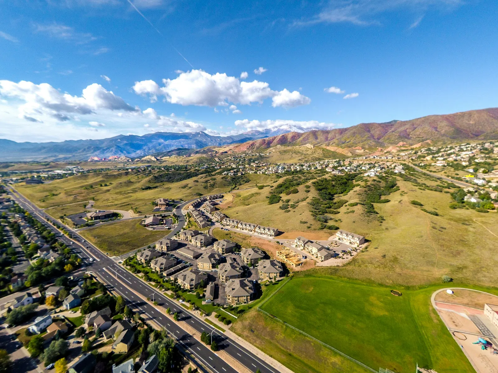 Aerial view of suburban homes, roads, and green fields with mountains in the background under a blue sky.