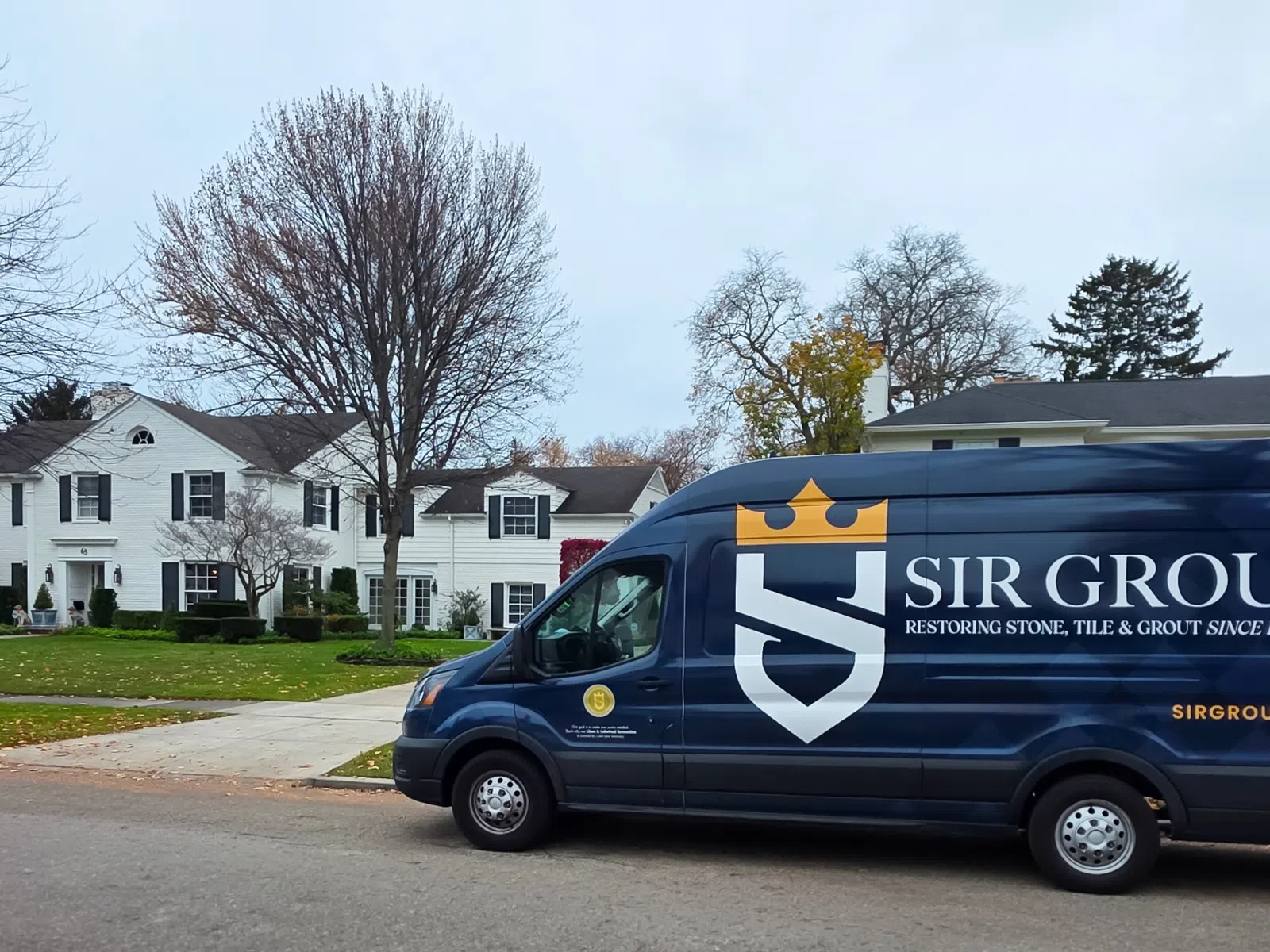 Dark blue Sir Grout van parked on a residential street in front of white houses with autumn trees and lawns.
