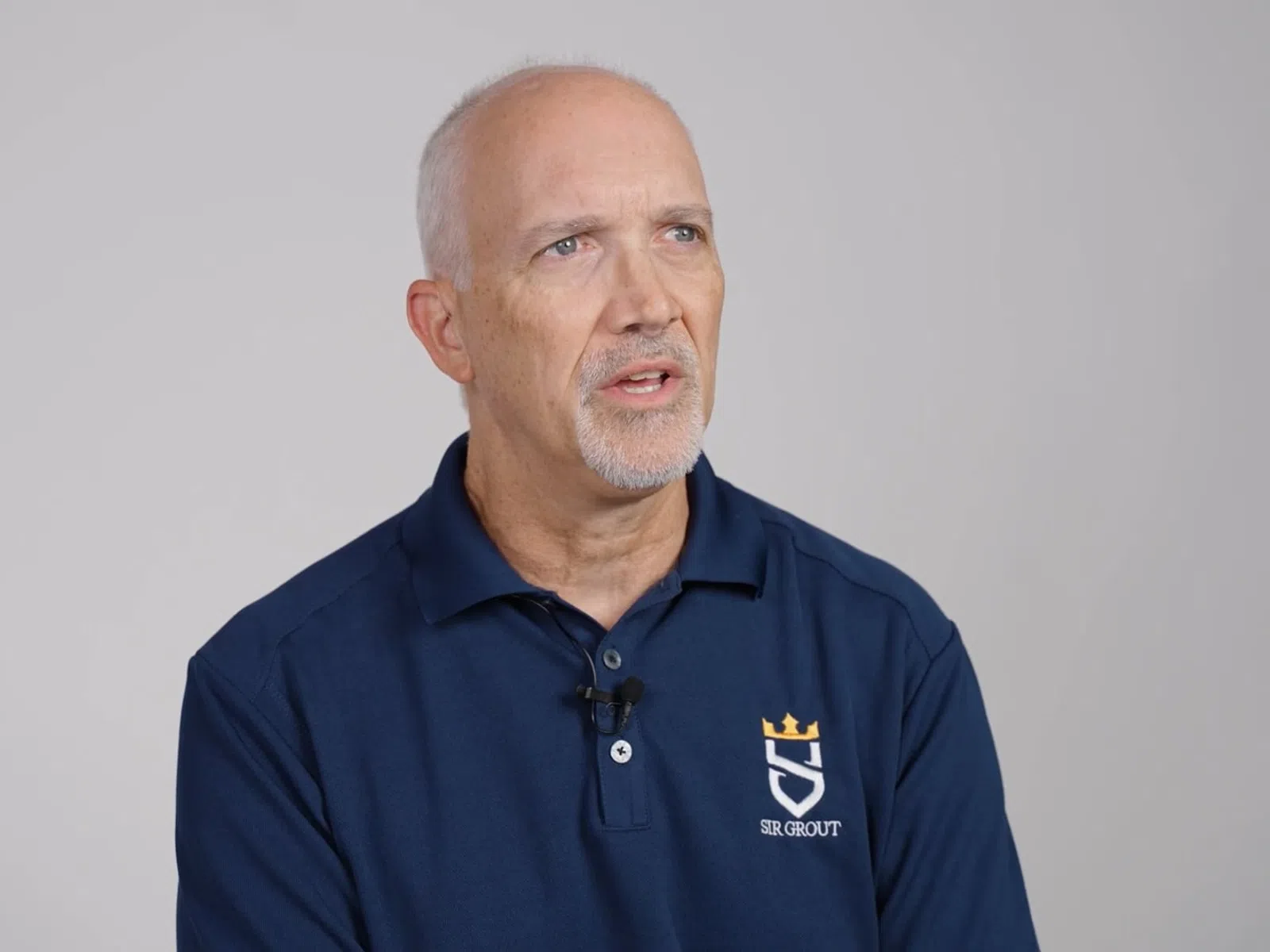 Mature man with gray hair wearing a navy blue Sir Grout polo shirt speaking against a plain gray background
