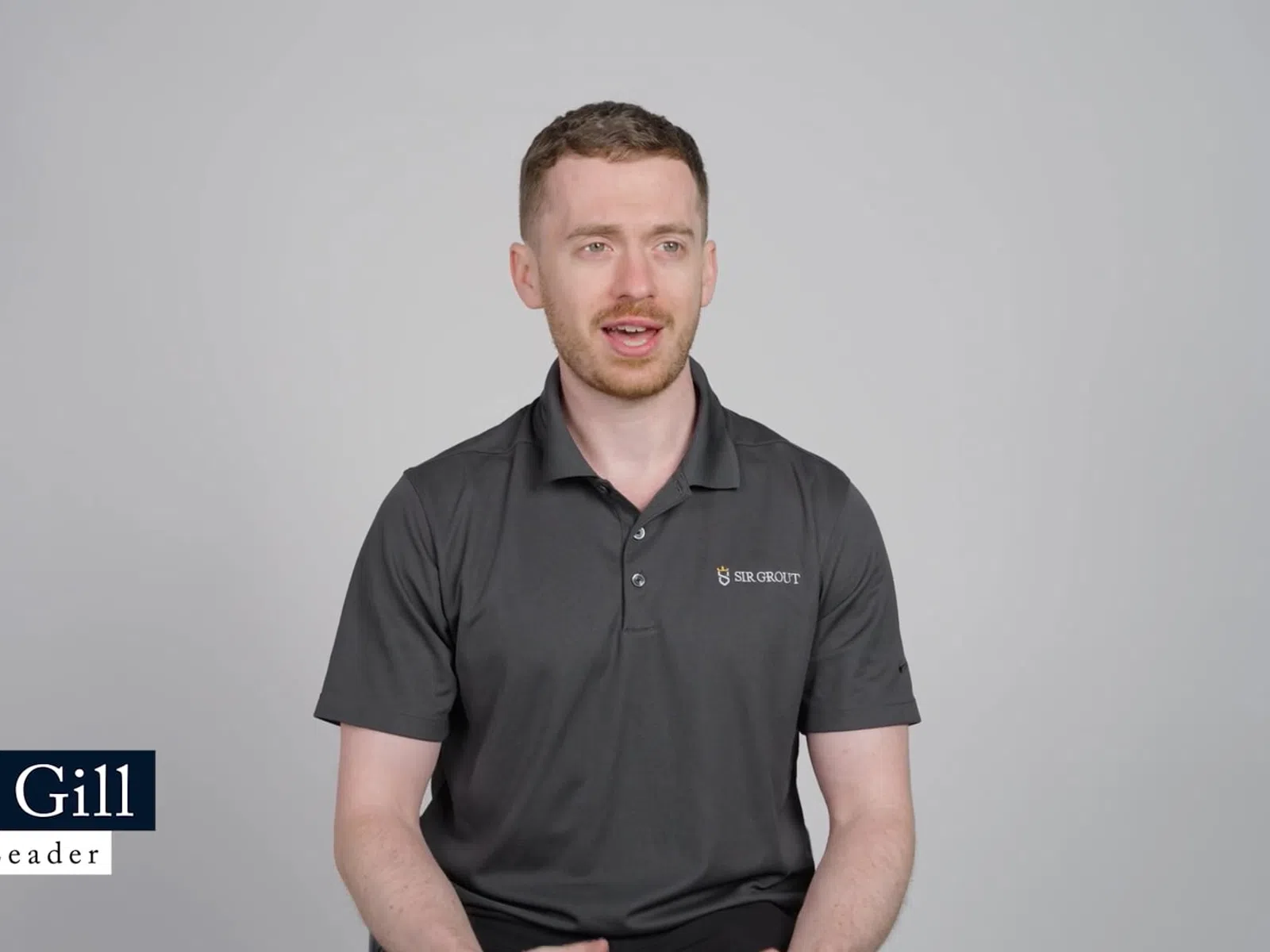Man in black polo shirt speaking against a plain gray background with name and title graphic overlay.