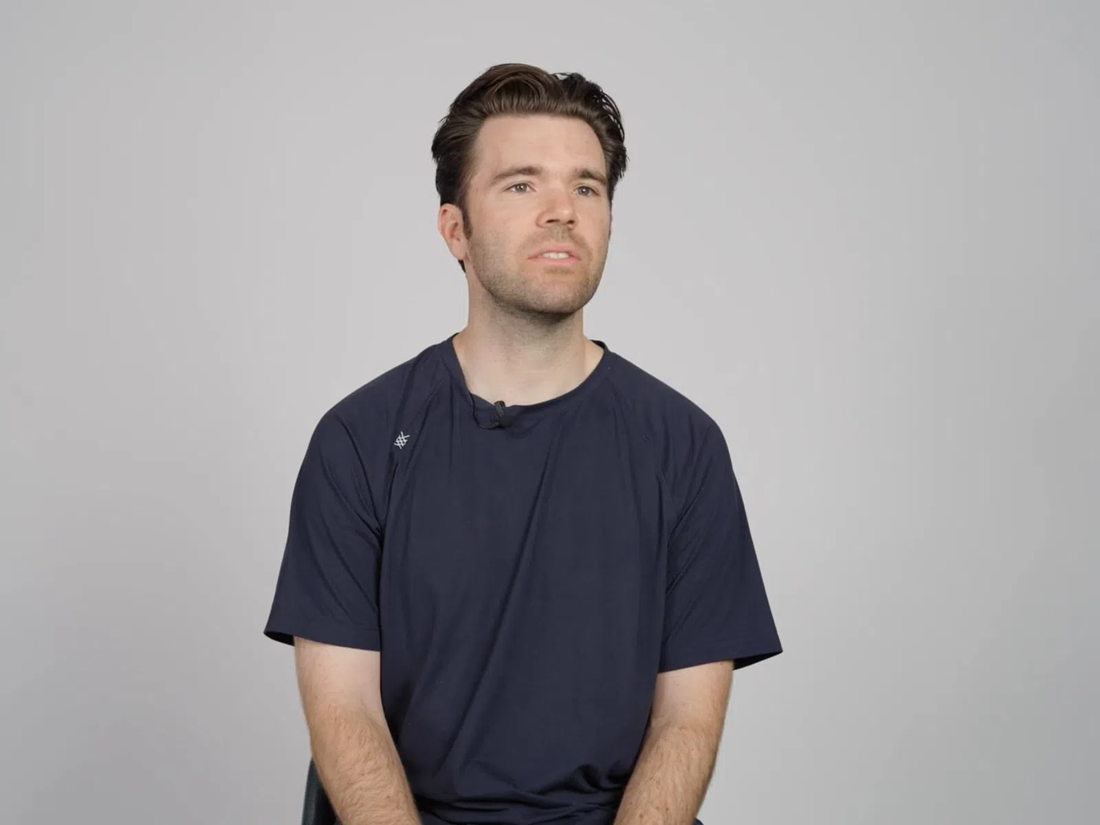 Young man with dark hair wearing a navy blue t-shirt sitting against a plain light gray background.