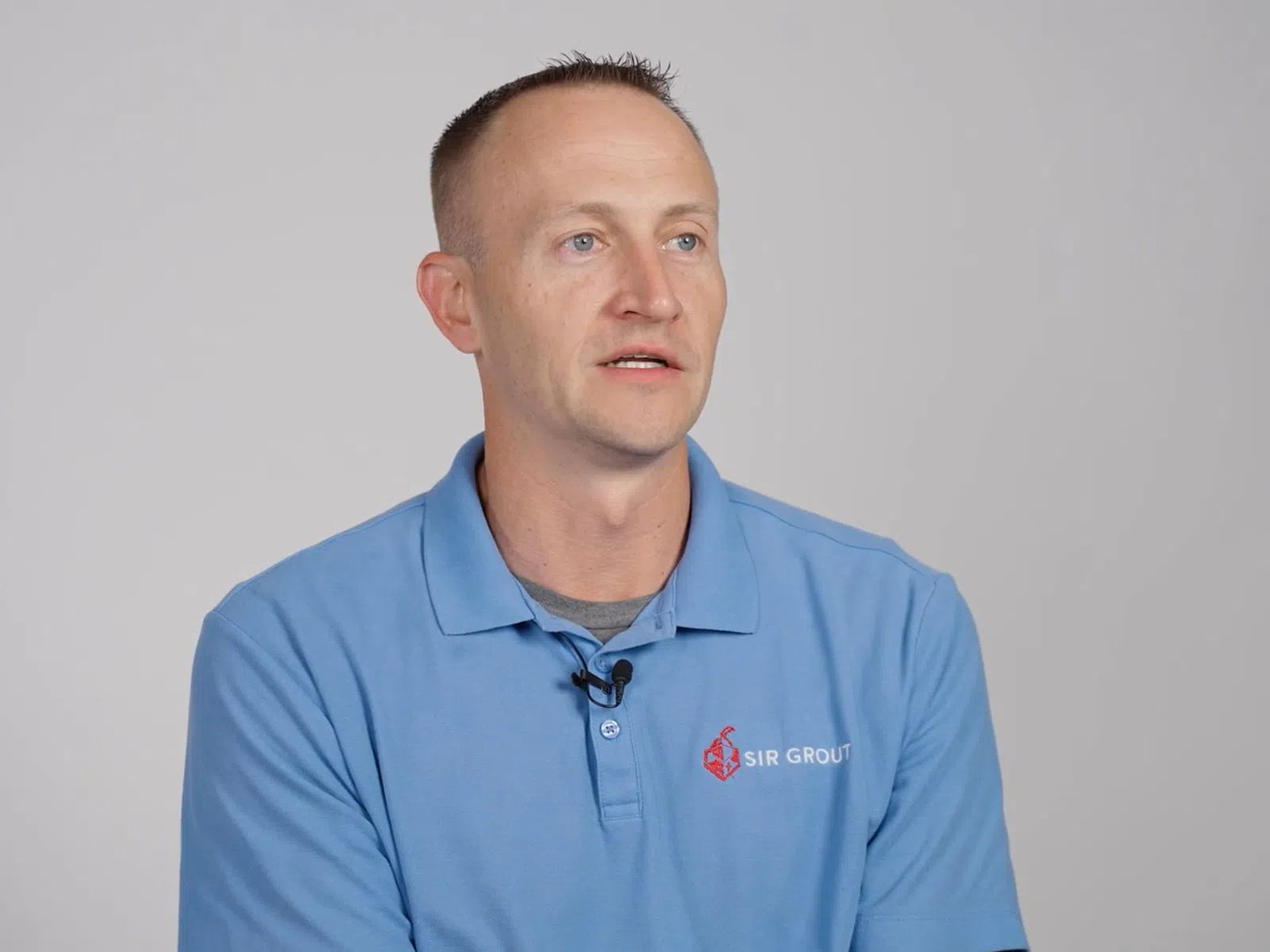 Man in blue Sir Grout polo shirt speaking against plain light gray background in interview setting