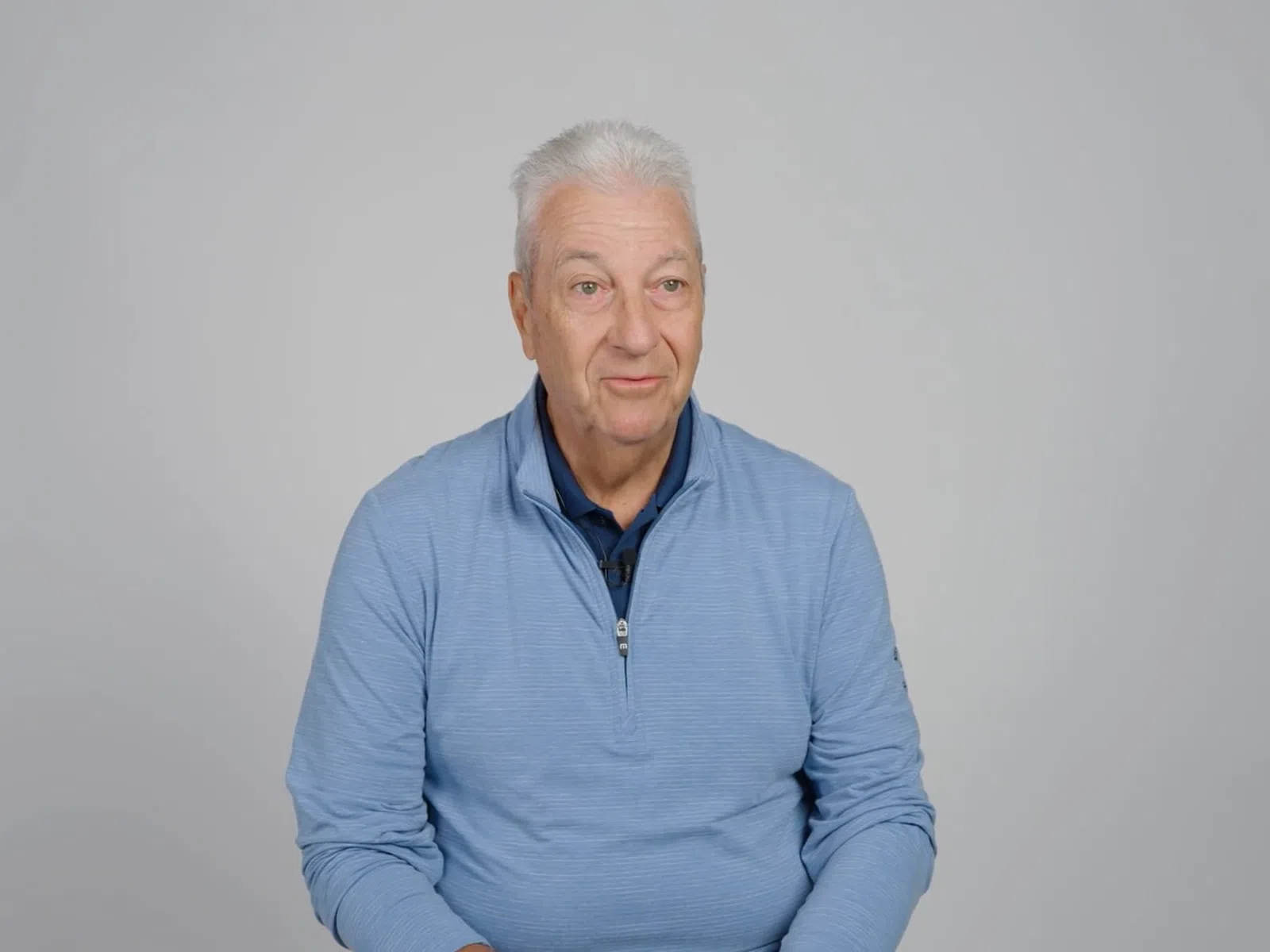 Senior man with white hair wearing a blue quarter-zip sweater sitting against a plain gray background.