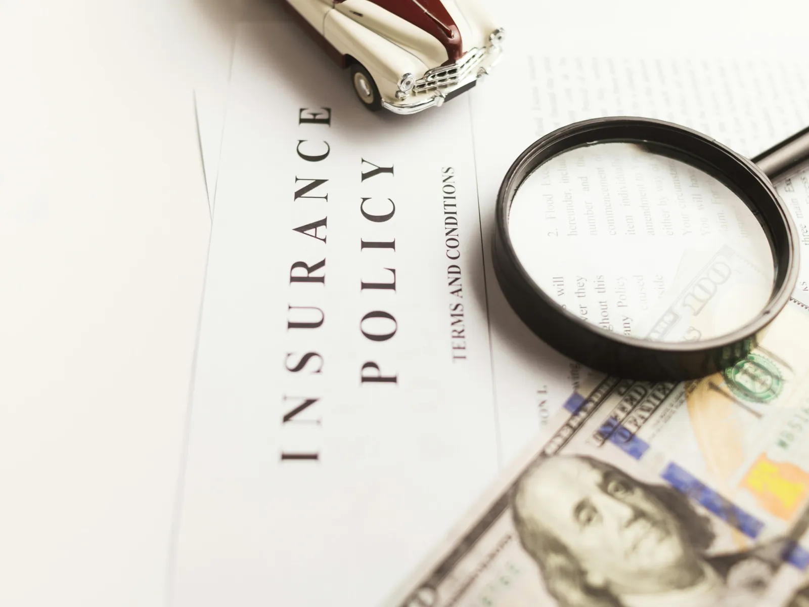 Magnifying glass over insurance policy document with hundred-dollar bill and toy car on white surface