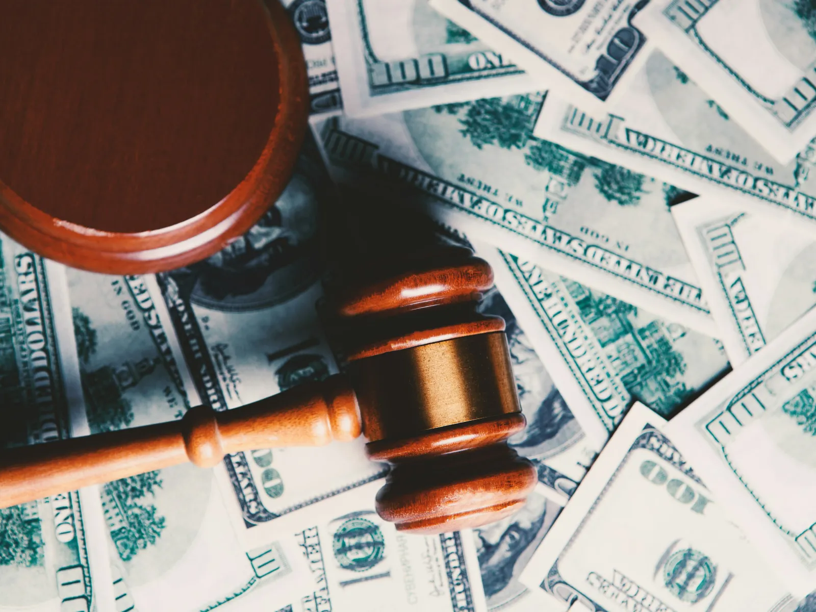 Wooden judge gavel with sound block on scattered US hundred dollar bills representing legal fines or financial judgment.