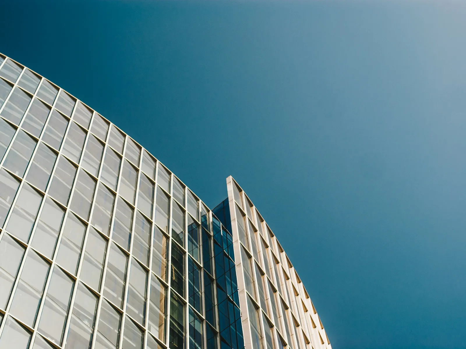Curved modern glass building facade against clear blue sky with reflective windows and metal framework.