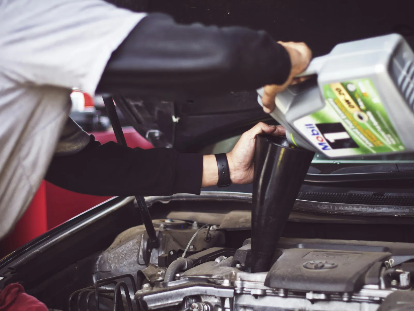 Person pouring motor oil into a car engine using a black funnel under the open hood of the vehicle.