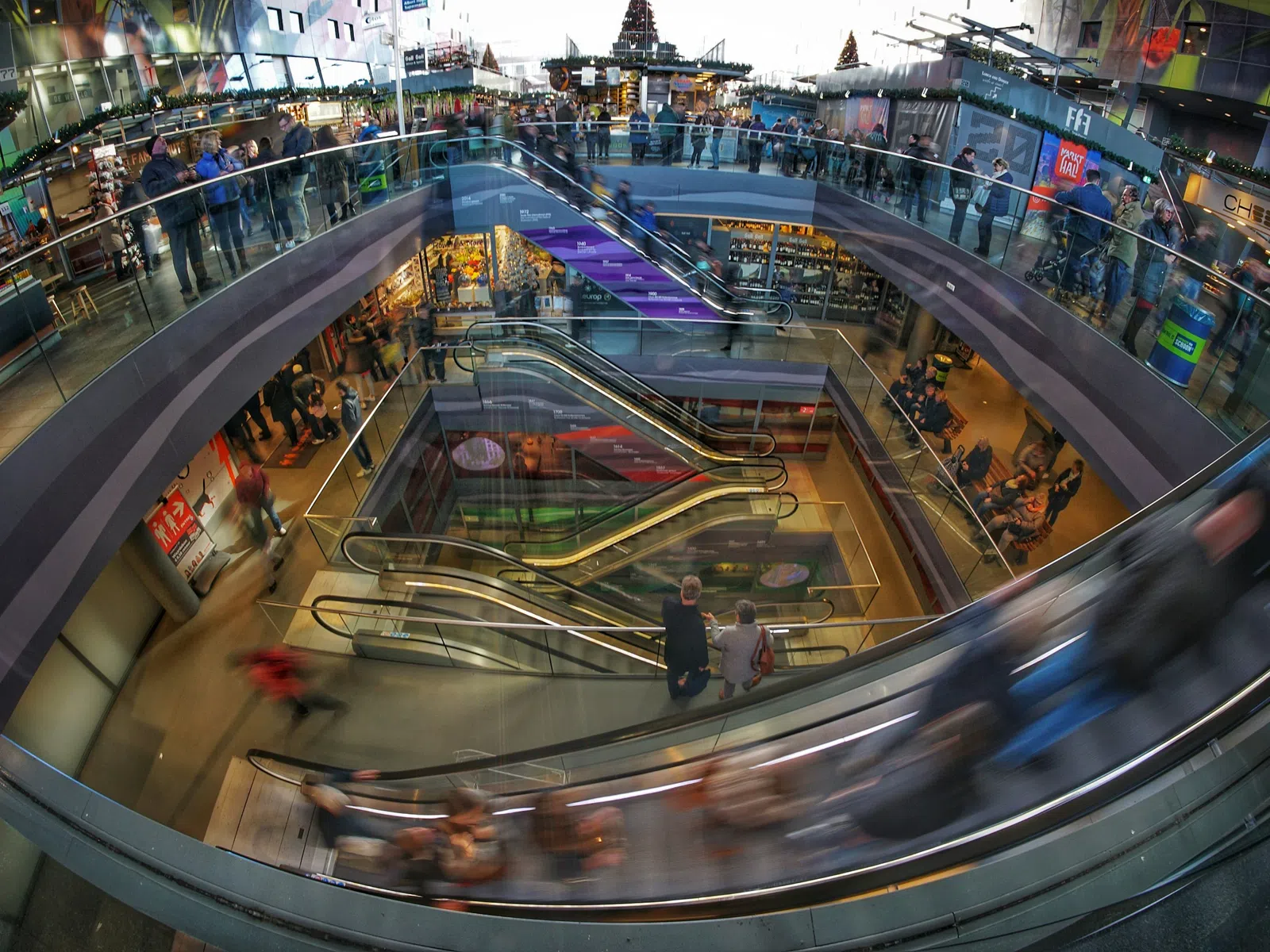 Busy modern multi-level shopping mall with escalators and people moving between floors.