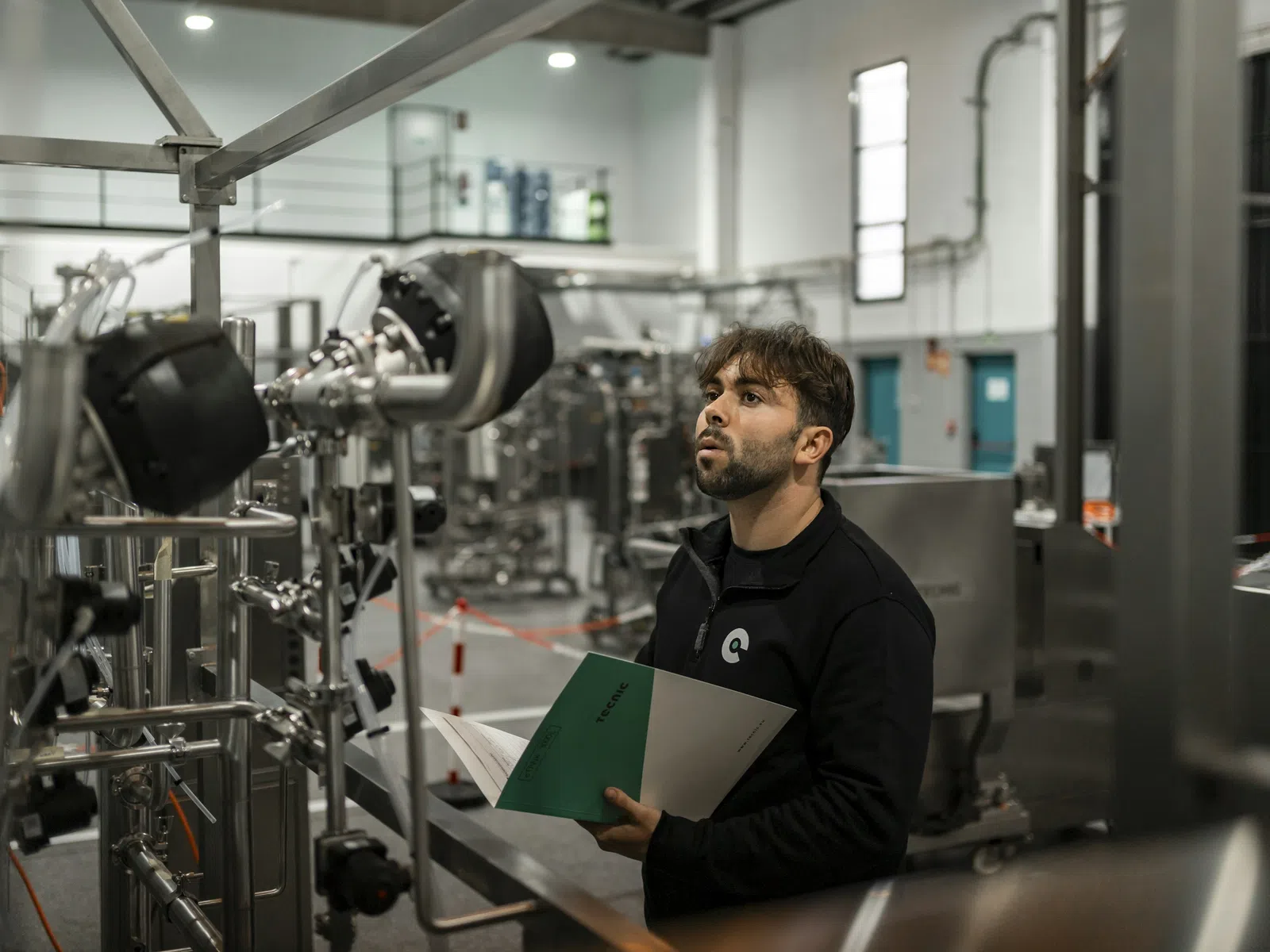 Man in black jacket inspecting machinery in a modern industrial facility holding a green and white clipboard.