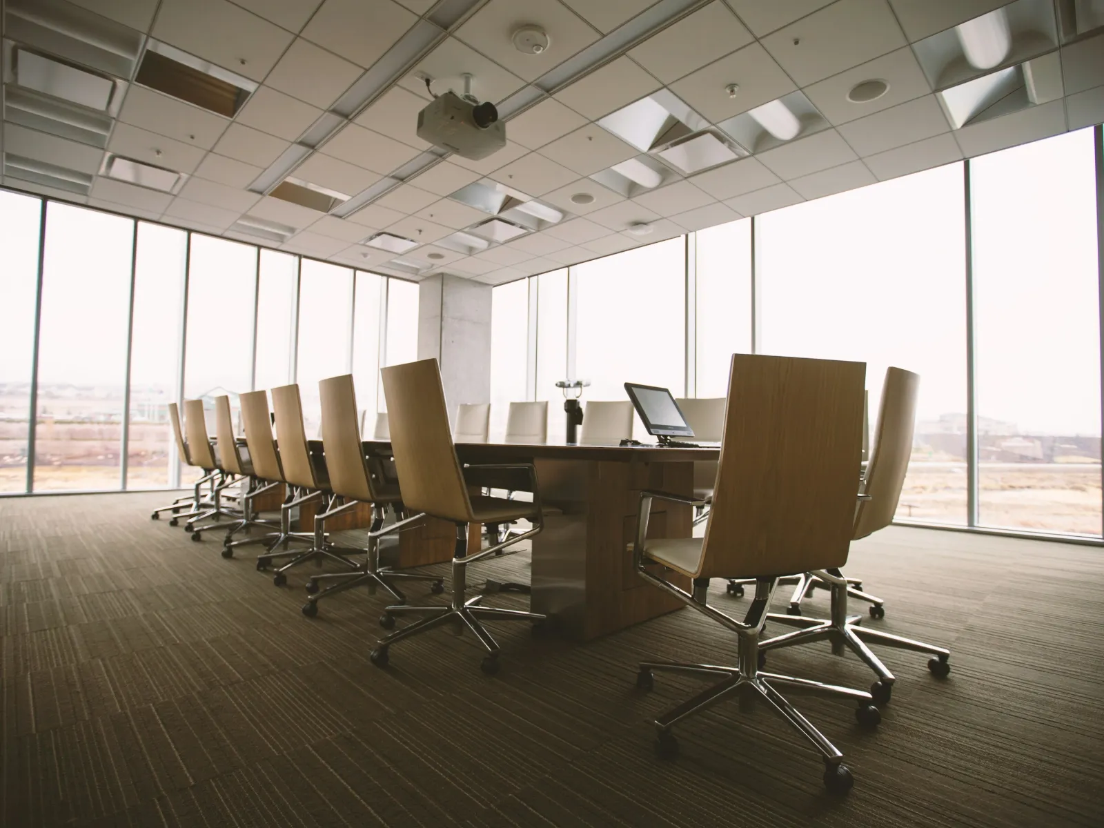 Modern empty conference room with long wooden table, swivel chairs, laptop, and large windows overlooking cityscape.