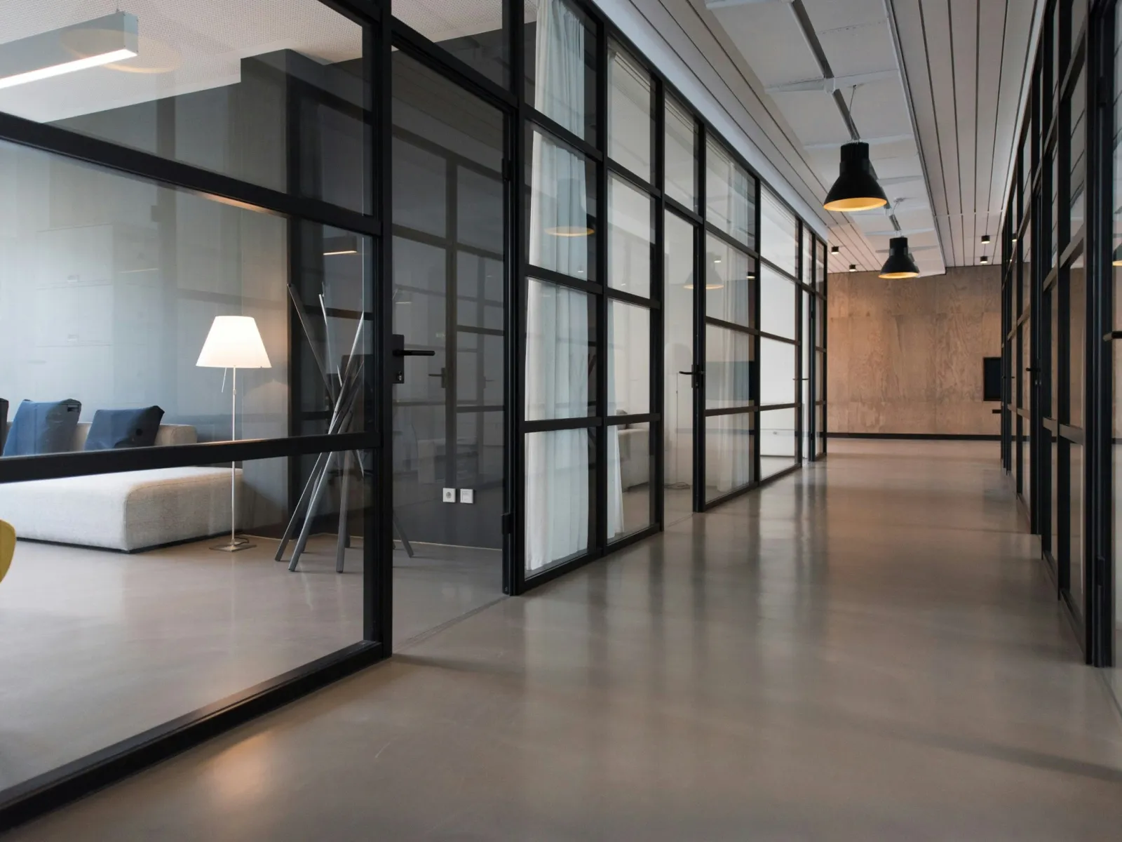 Modern office hallway with glass-walled rooms, concrete floors, pendant lights, and minimalistic furniture.