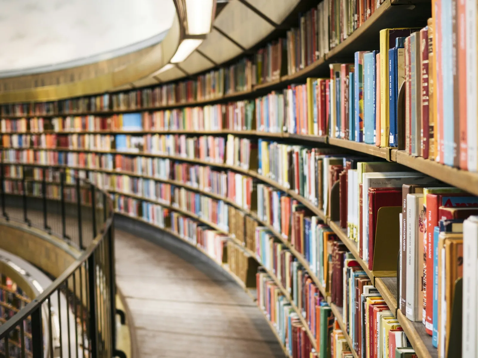 Curved wooden bookshelf filled with colorful books lining a winding library aisle with metal railing.