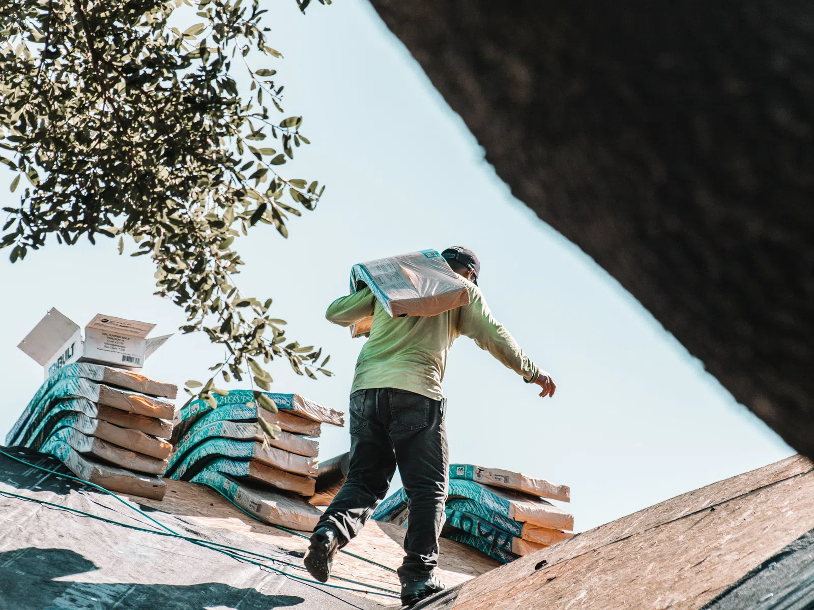 Construction worker carrying a heavy bag on a roof next to stacked bags under a clear sky.