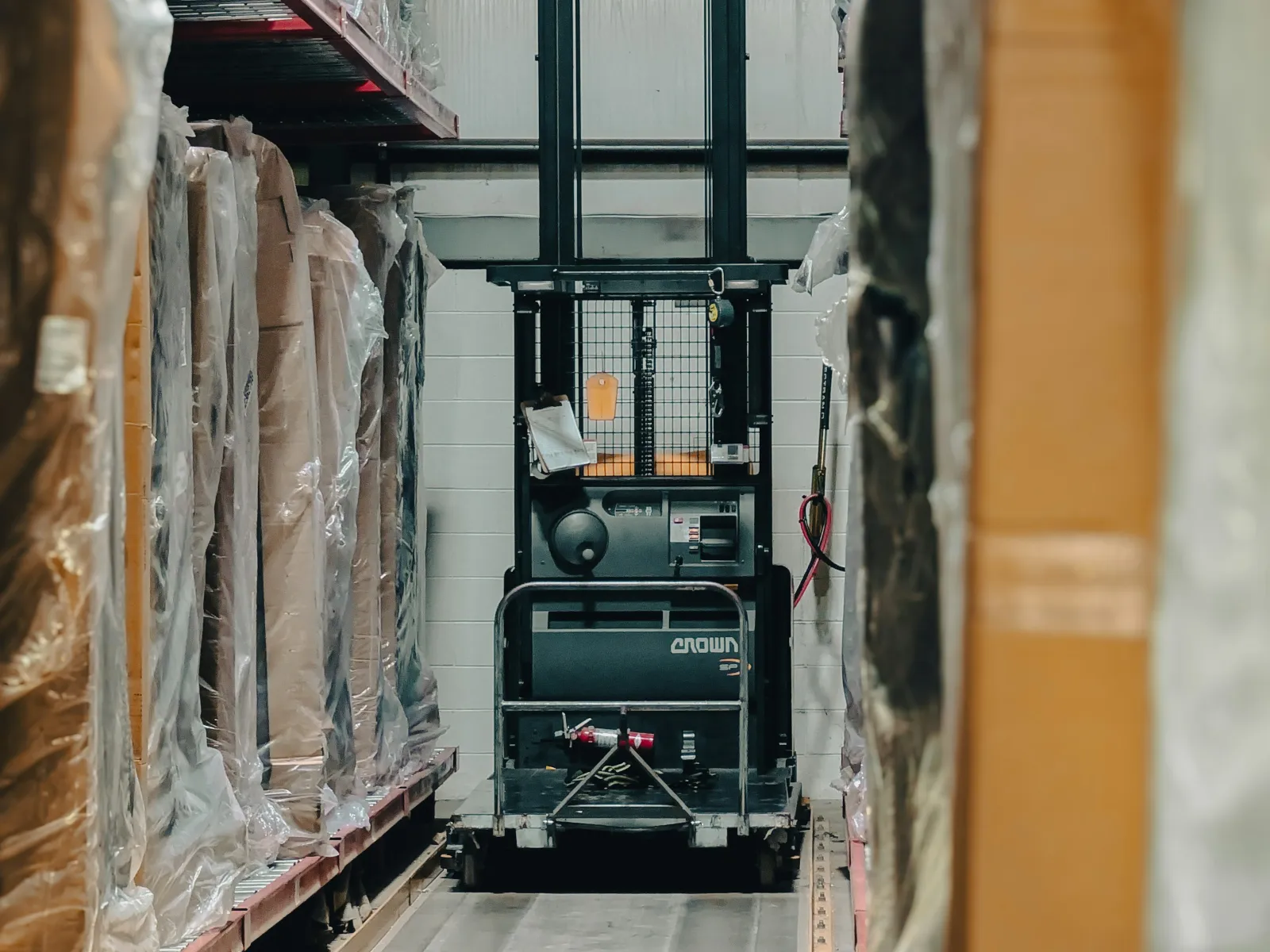 Forklift moving through narrow warehouse aisle between shelves stacked with wrapped goods and boxes.