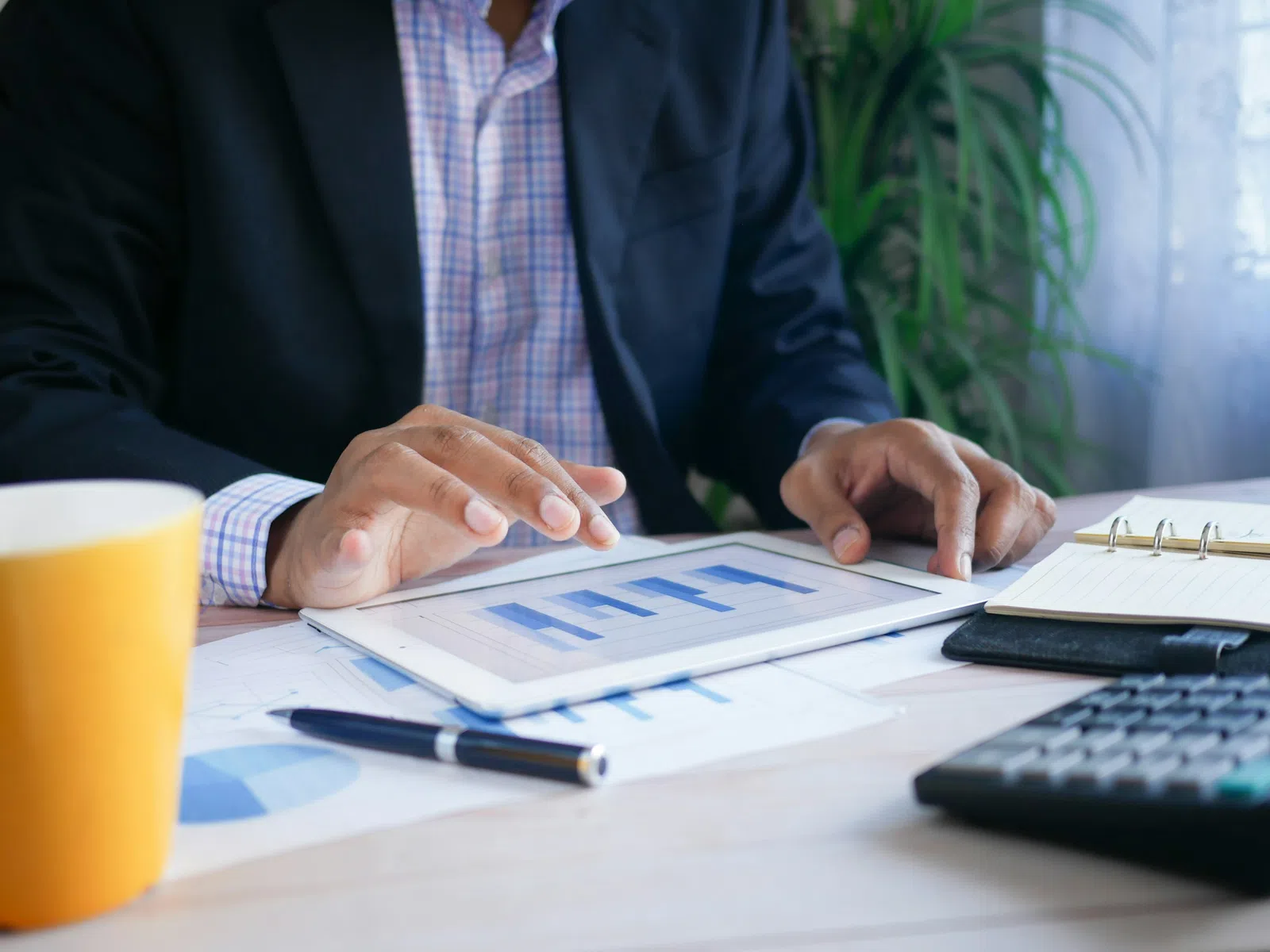 Businessman analyzing blue bar charts on a tablet with calculator, notebook, pen, and yellow cup on desk