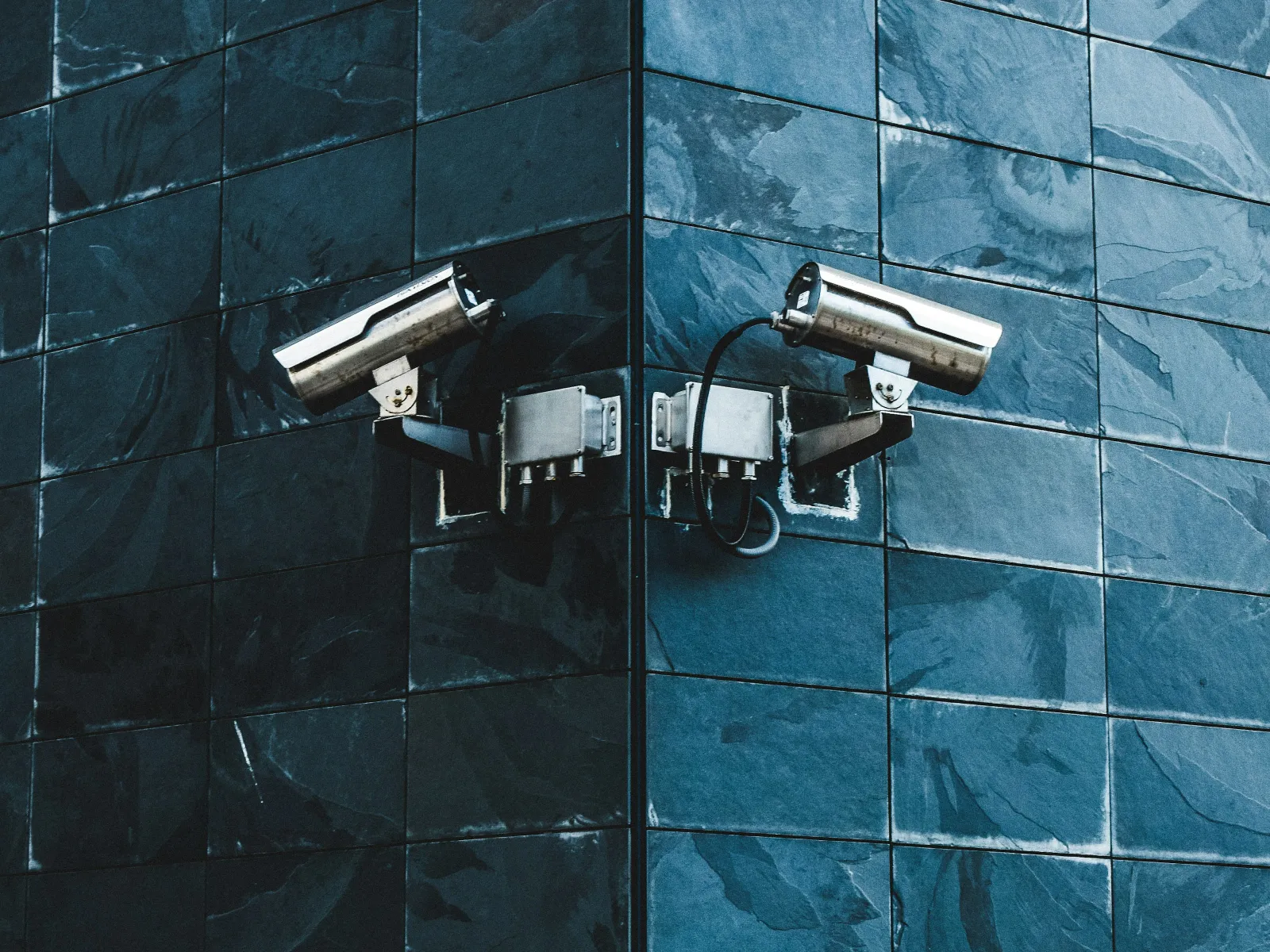 Two security cameras mounted on the corner of a blue tiled building wall monitoring opposite directions.