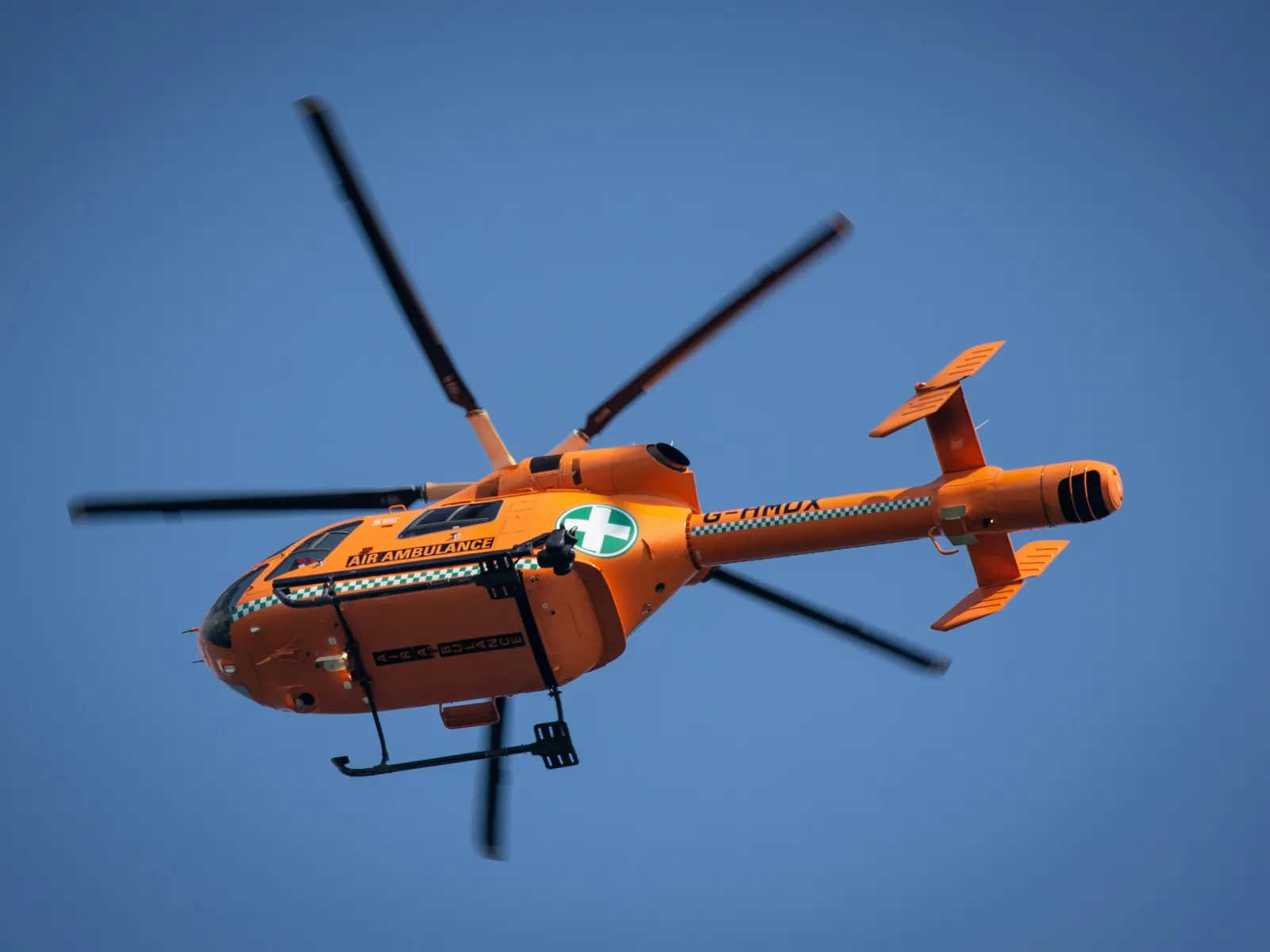 Orange air ambulance helicopter with rotating blades flying against clear blue sky background.