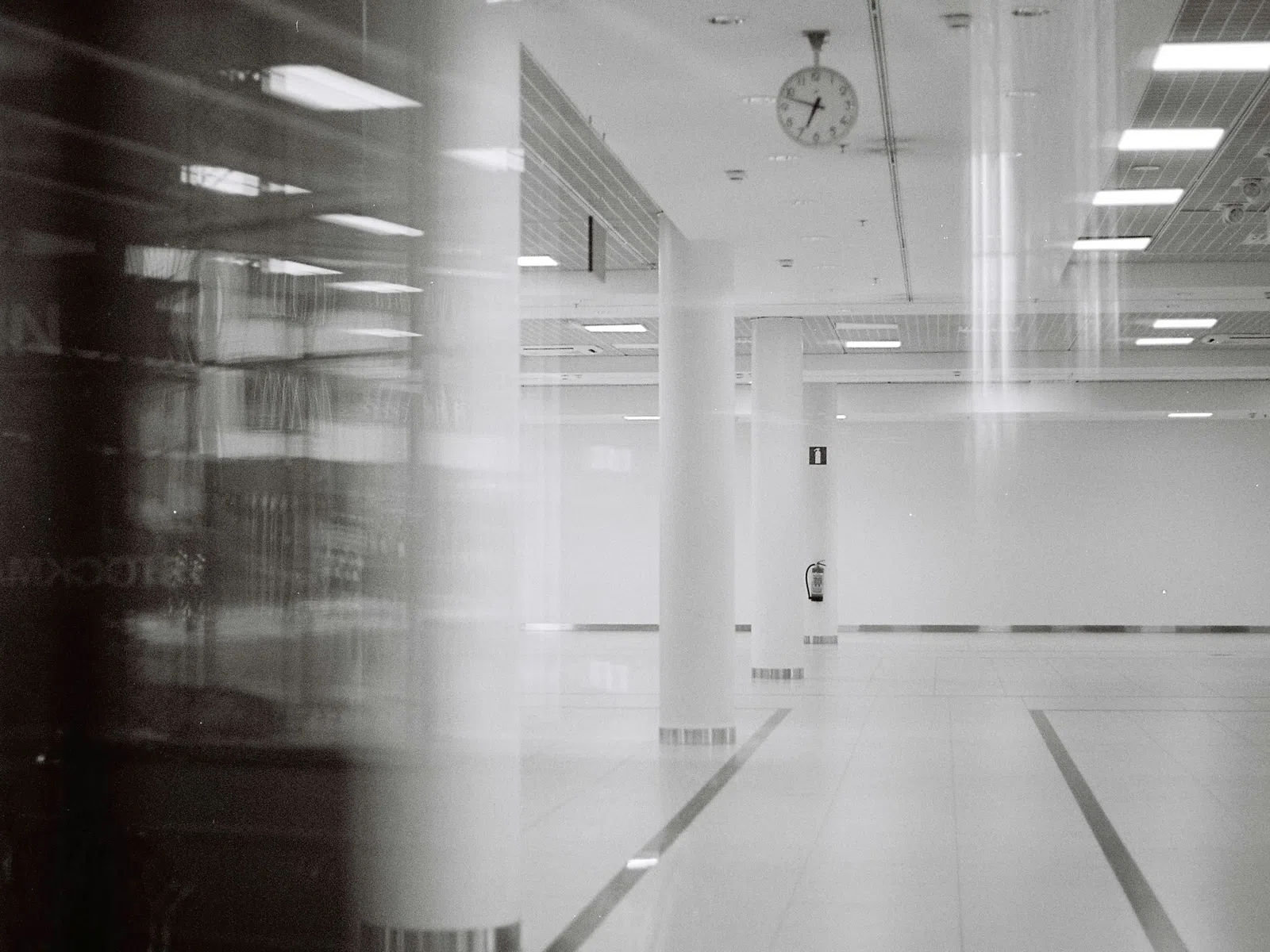 Empty modern hallway with white columns, tiled floor, ceiling lights, and a clock on the ceiling showing 2:55.