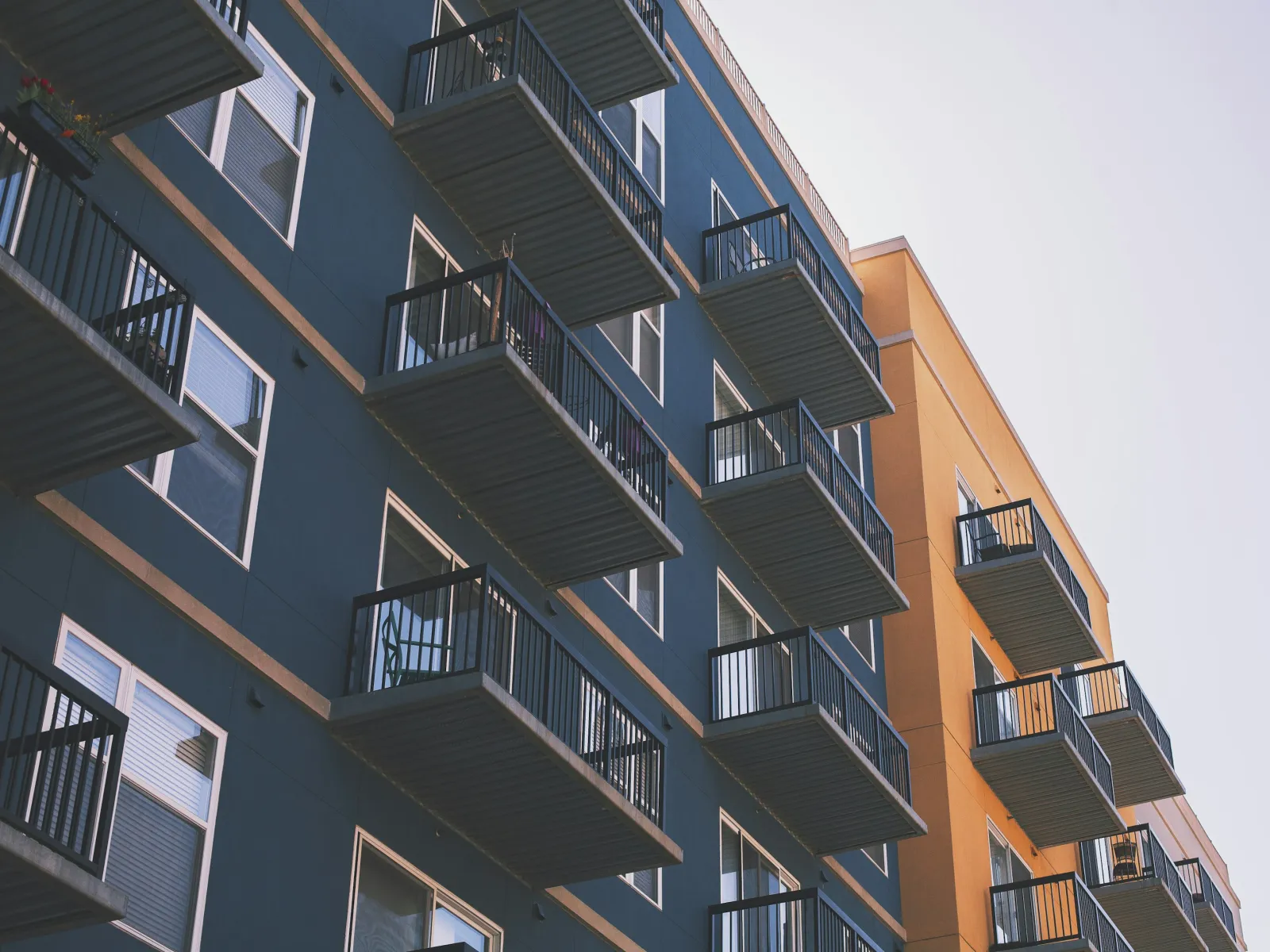Modern apartment building with black metal balconies against blue and yellow exterior walls under clear sky.