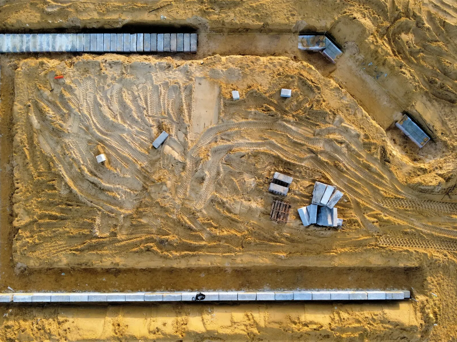 Aerial view of a sandy construction site with concrete blocks and vehicle tire tracks forming a rectangular foundation.