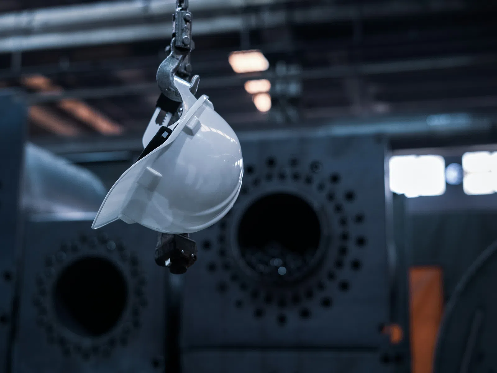 White safety helmet hanging from a hook in an industrial factory with machinery in the background.