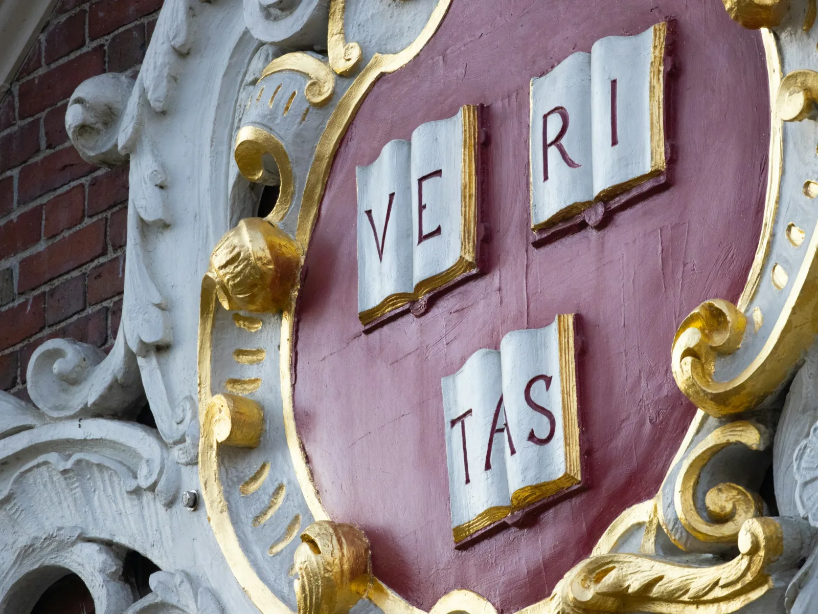 Close-up of Harvard University shield with red background, gold trim, and three open books spelling Veritas.