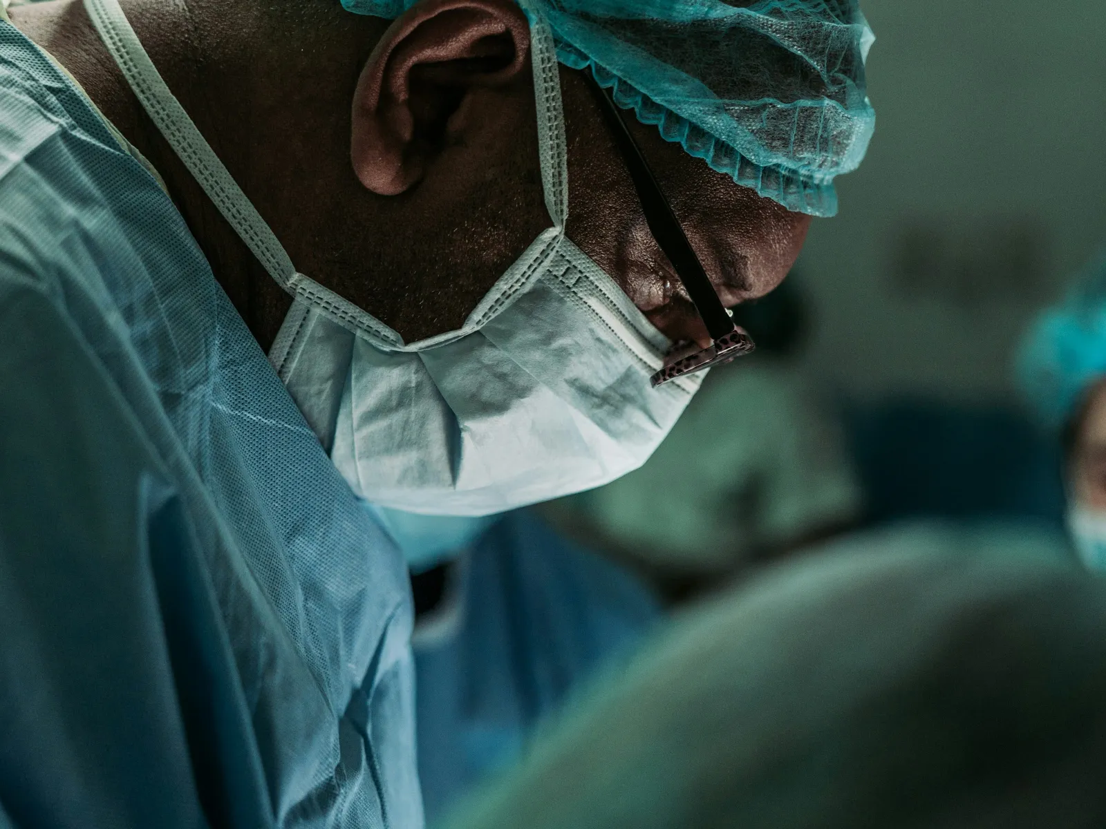Surgeon wearing surgical mask, cap, and gown focused on operation in sterile operating room environment.