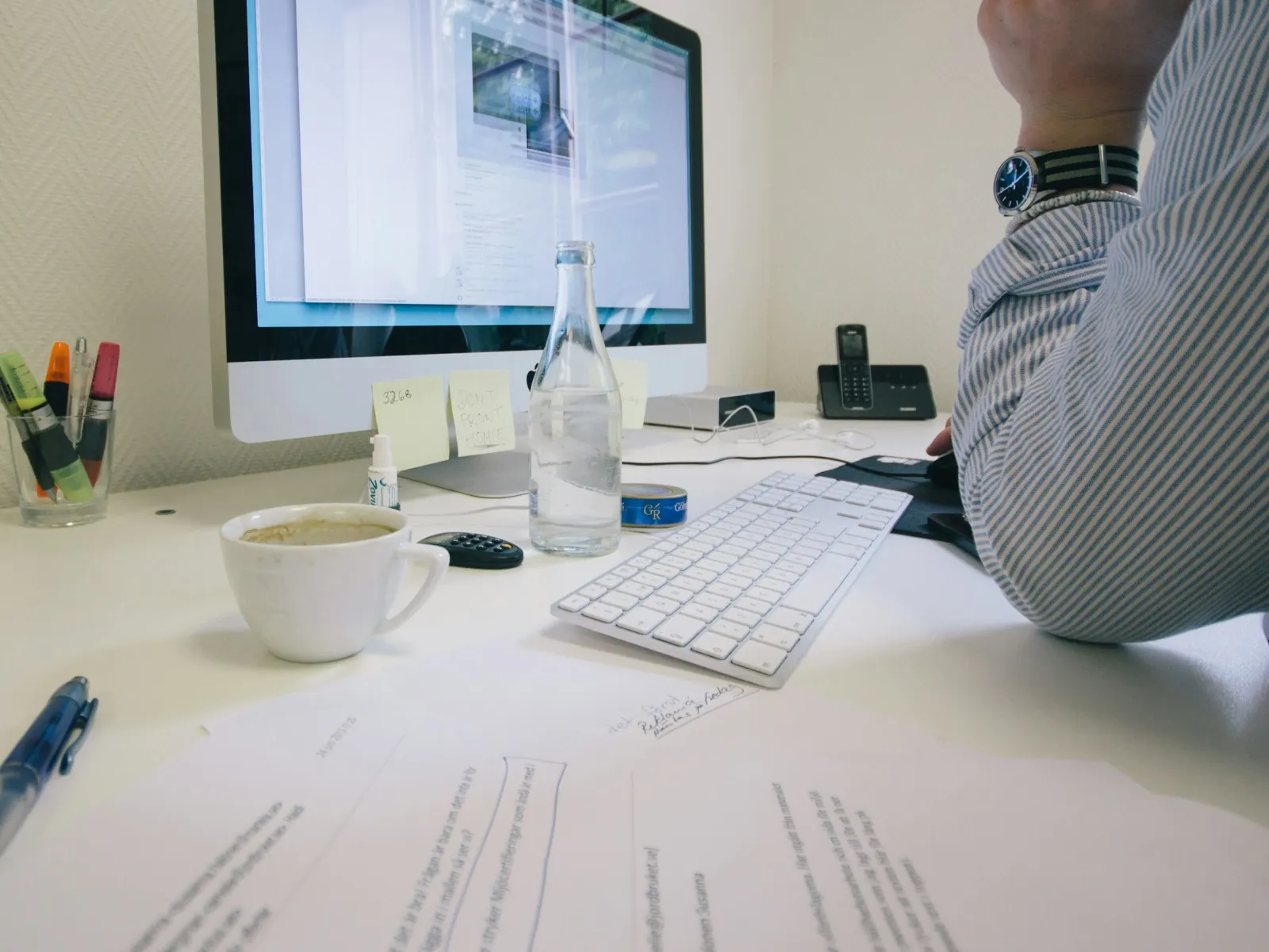 Workspace with a computer, coffee cup, bottled water, documents, pens, and a person in striped shirt at desk.
